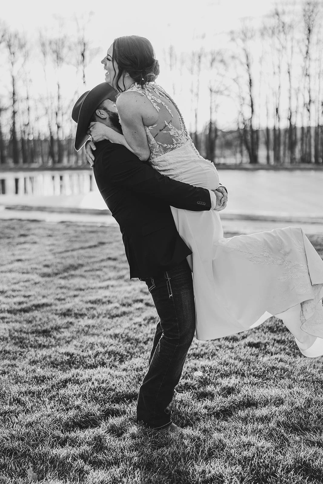 A black-and-white photo of a man lifting a woman in a wedding dress outdoors by a lake, with trees in the background.