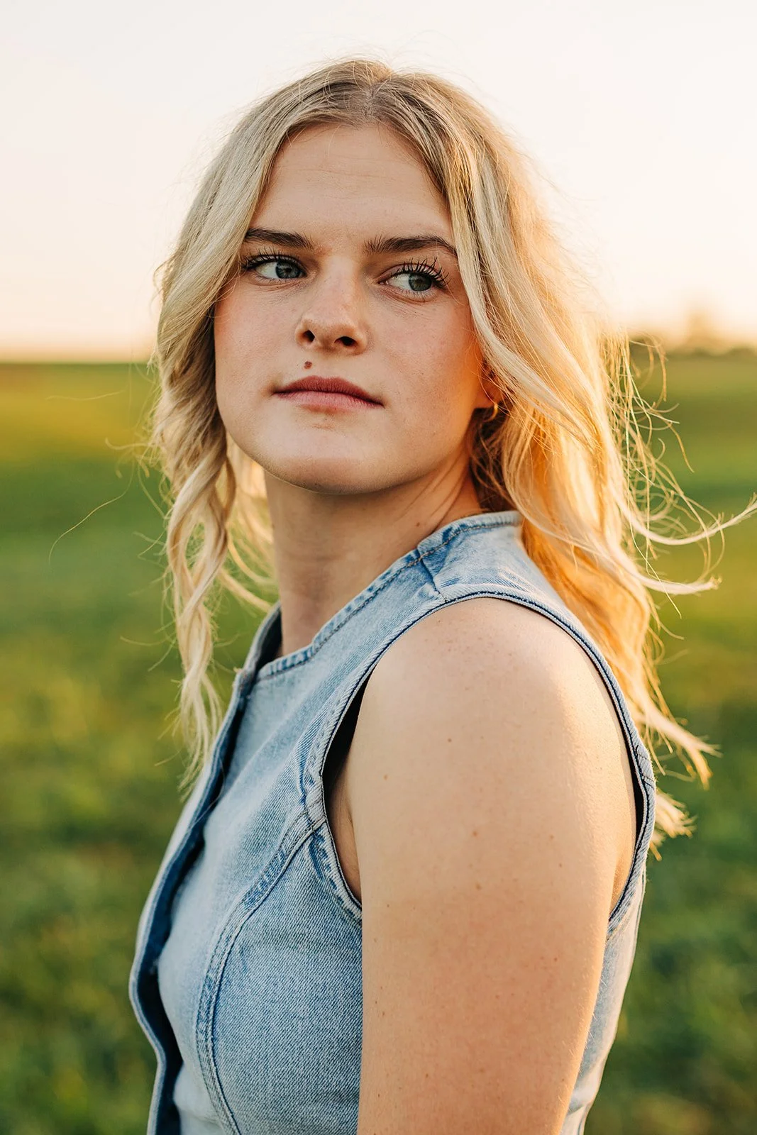 A young woman with blonde wavy hair and blue eyes, wearing a denim sleeveless top, standing outdoors in a green field during sunset.