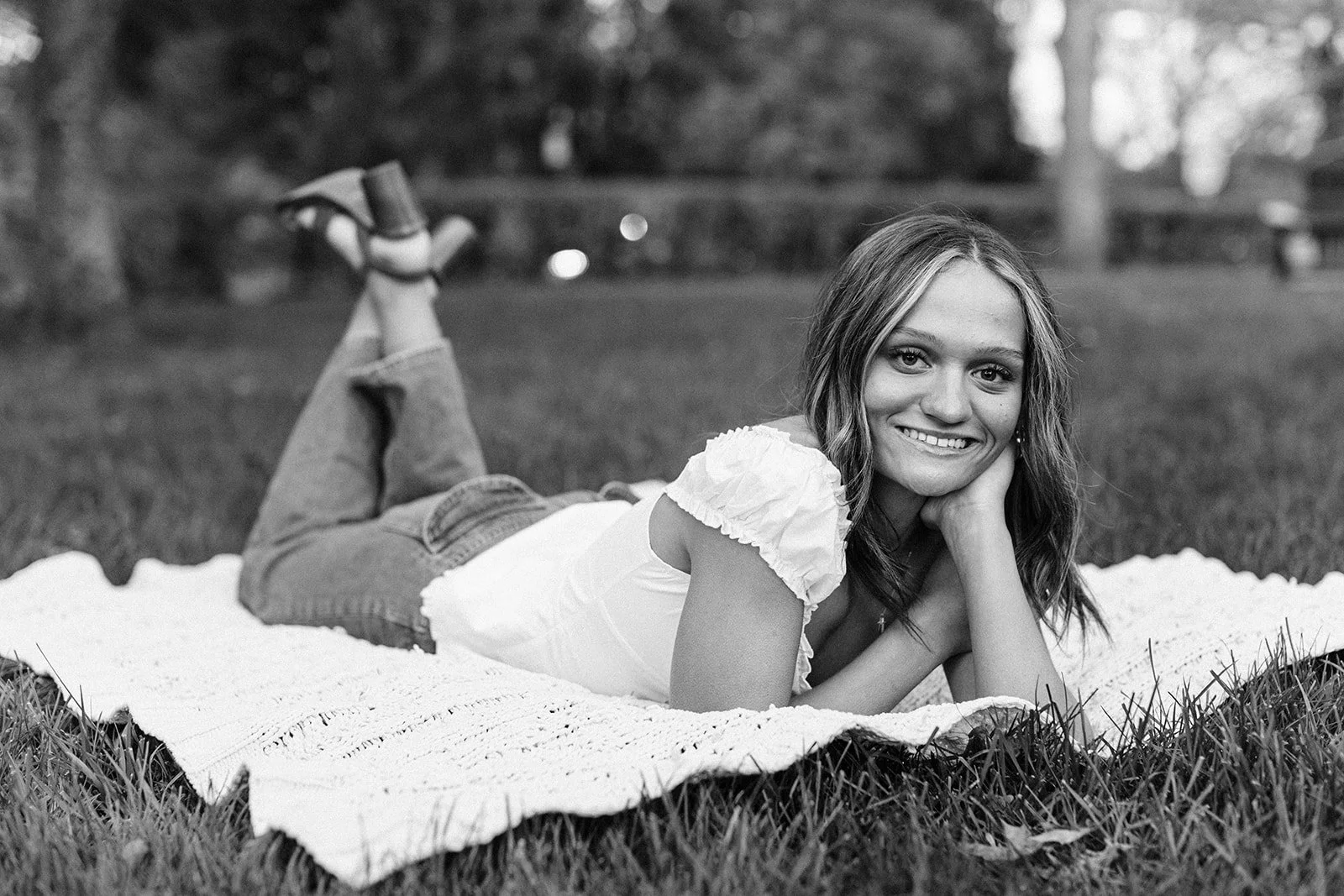 A young woman lying on her stomach on a blanket in a park, smiling, with her head resting on her hand. She has long wavy hair and is wearing a short-sleeved top and jeans. The background shows trees and a grassy area.