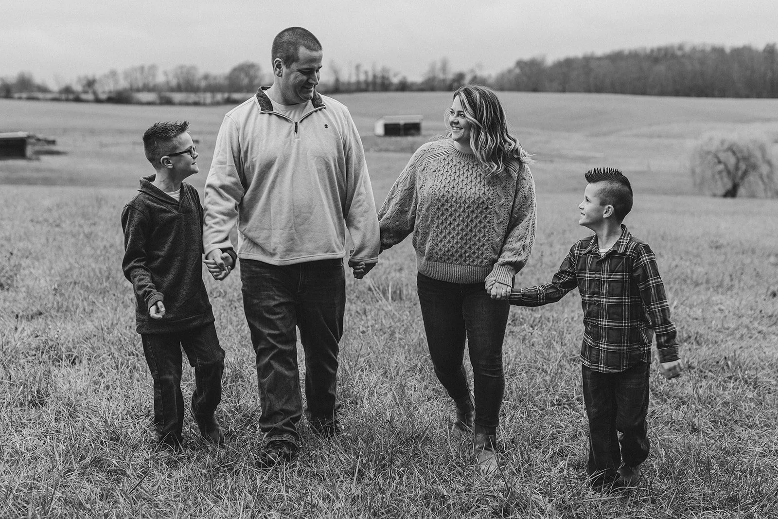 A black and white photo of a family of four walking hand in hand through a grassy field, smiling and looking at each other.