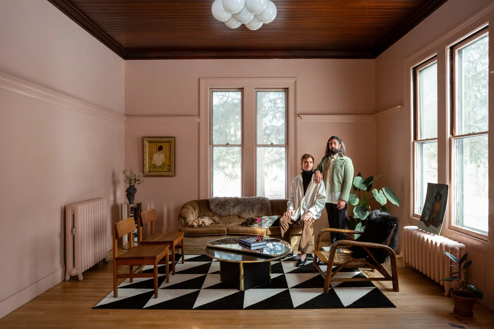 A living room with pink walls, wooden ceiling, large windows, and a black and white geometric patterned rug. Two men are posing in front of a tan-colored sofa with a dog laying on it. The room has chairs, a coffee table, a painting of a woman, a potted plant, and a round ceiling light fixture.