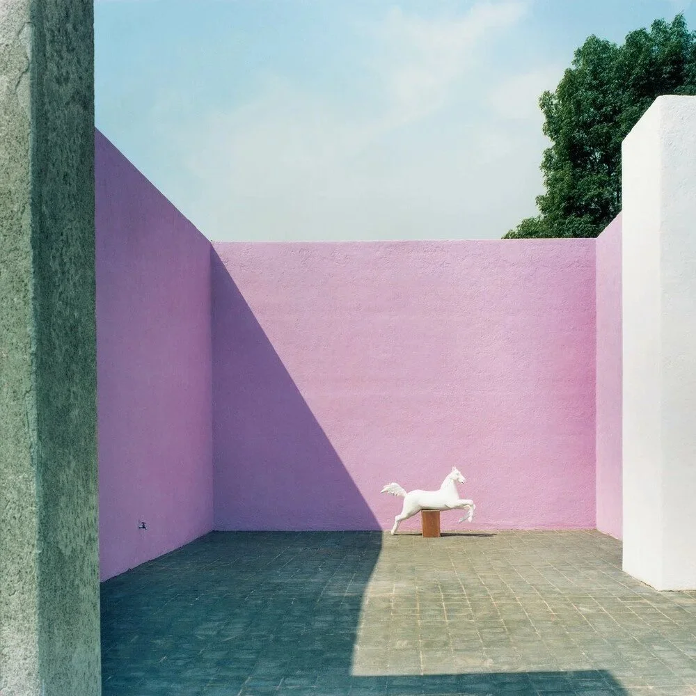 A minimalist courtyard with pink and white walls, a horse sculpture on a small pedestal, and a large shadow cast across the ground, with trees and a blue sky in the background.