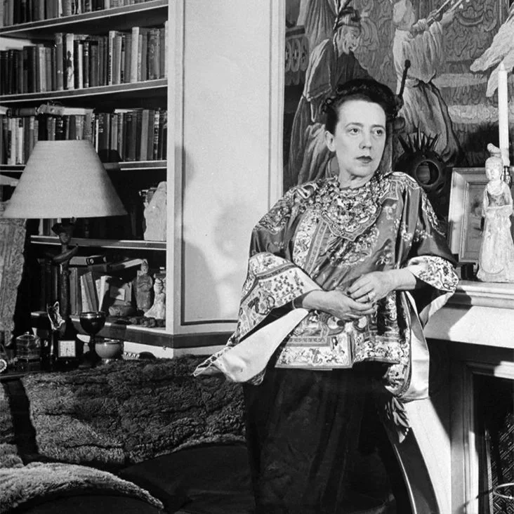A woman with dark hair styled up, wearing a patterned kimono, stands in a living room beside a fireplace with various decorative items and a large painting or wall art behind her. There are books on a bookshelf, a lamp, and a cozy couch.