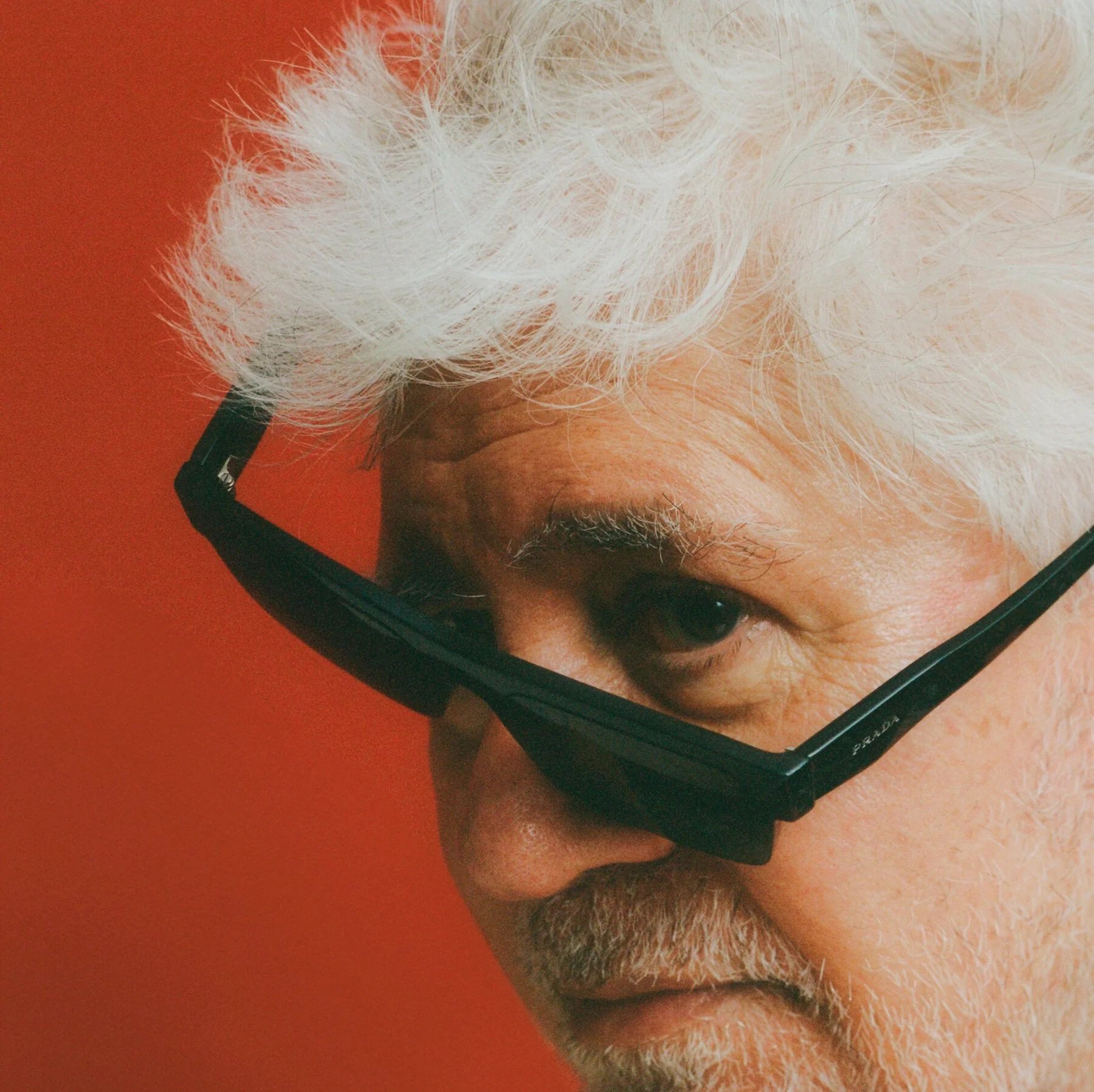 Close-up of a person's face with white hair, wearing glasses, looking down with a beard, on an orange background.