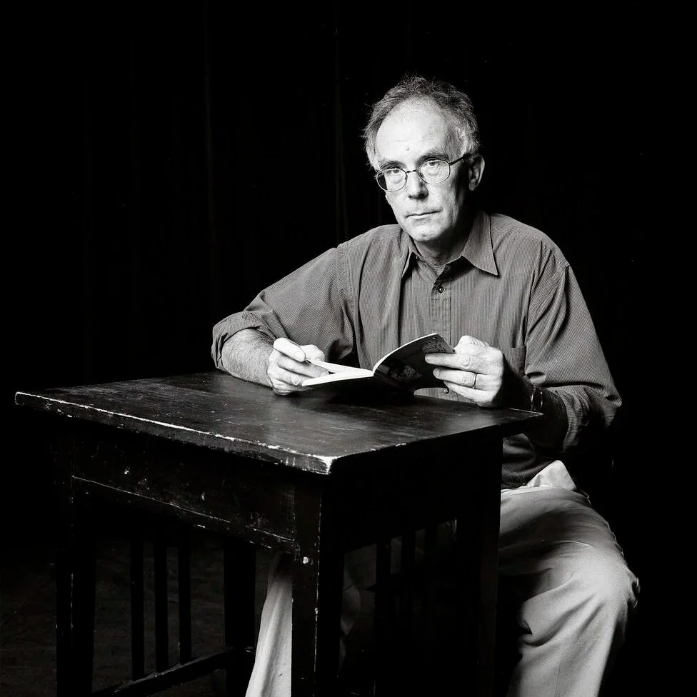 Black and white photo of a man sitting at a wooden table, holding a book and a pen, with a serious expression, against a dark background.