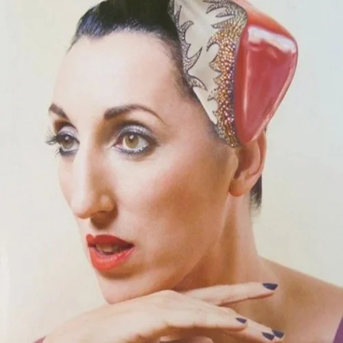 A woman with dark hair styled in a sleek updo, wearing bold makeup, and posing with her head resting on her hand. On her head, she has a partially visible decorative headpiece resembling a brain, with detailed patterns and textures.
