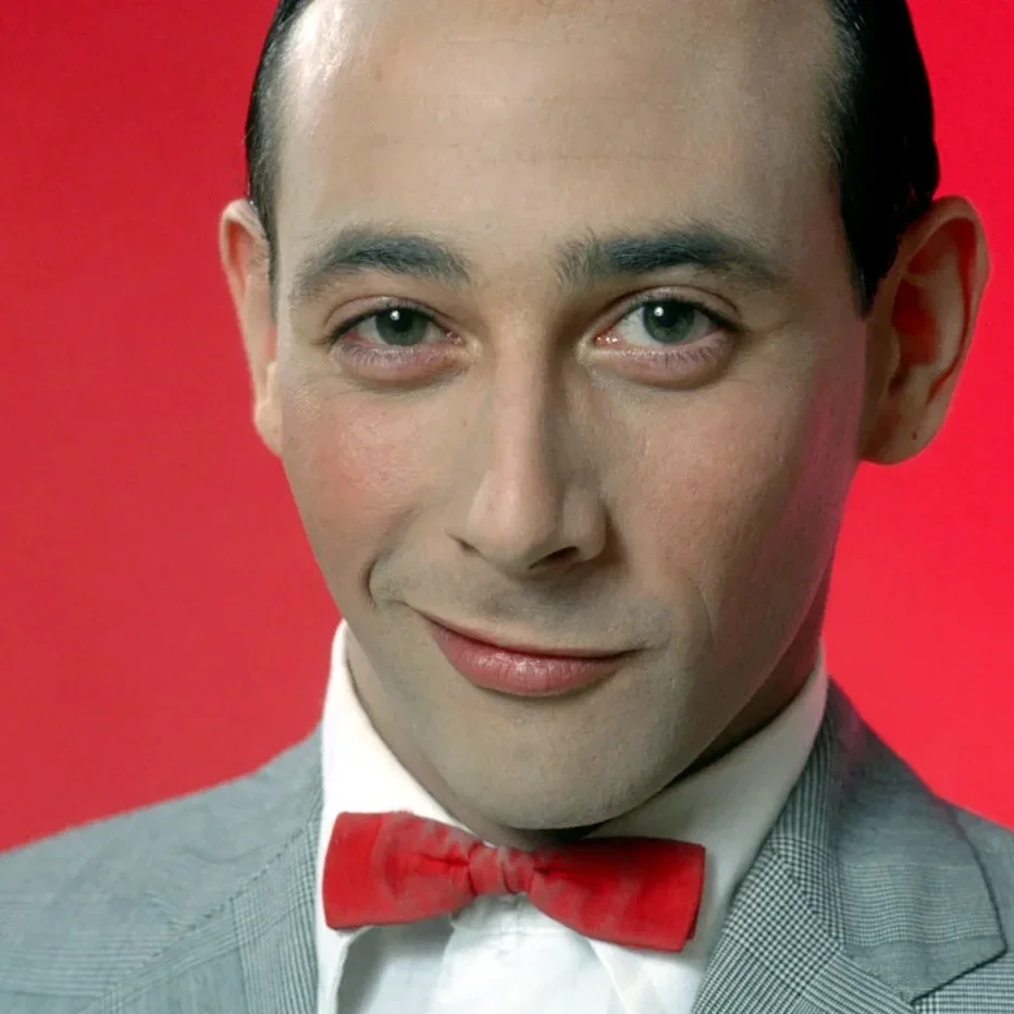 Close-up of a man with fair skin, slicked-back dark hair, wearing a gray suit, white shirt, and red bow tie, smiling with a confident expression against a red background.