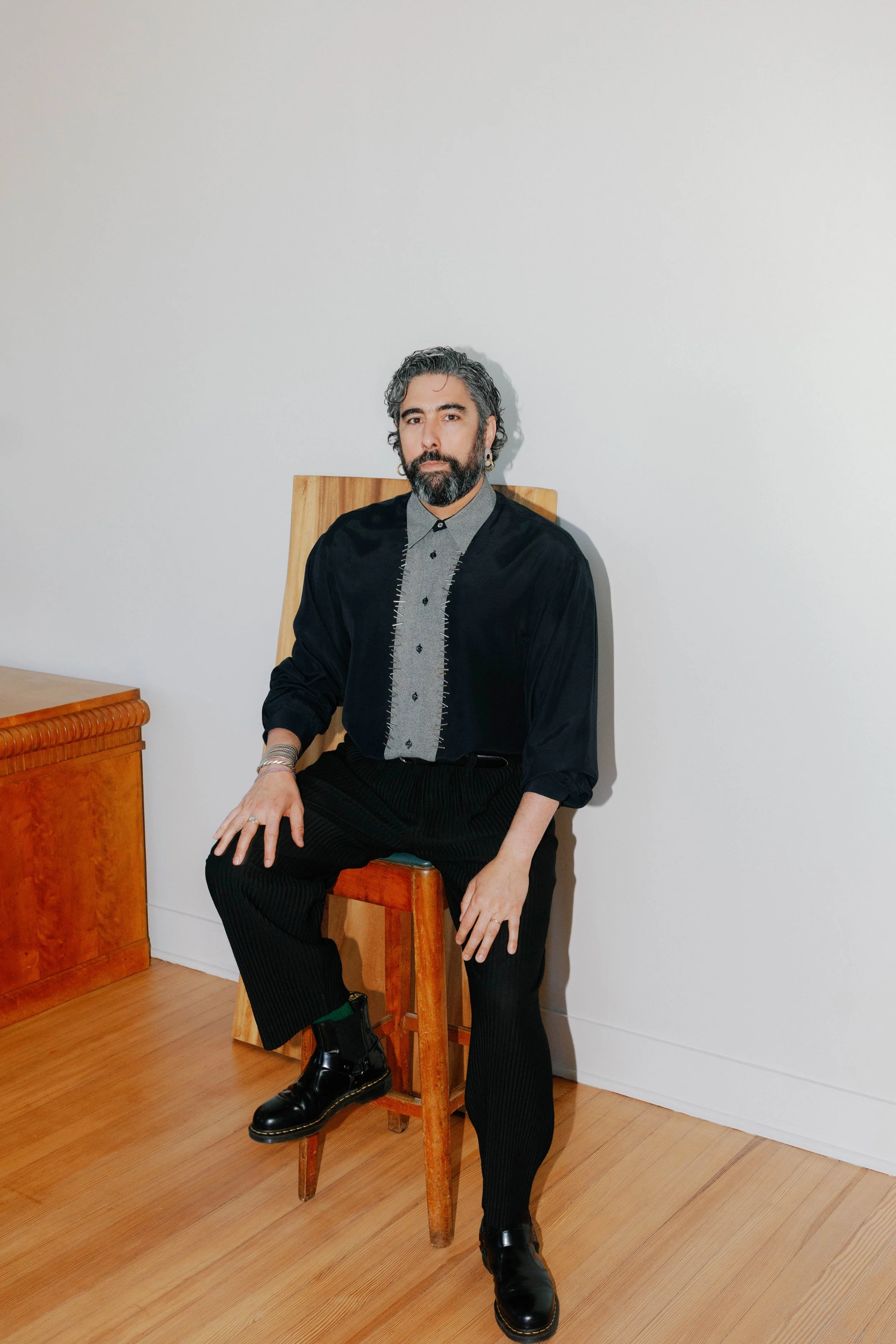 A man with a beard and curly gray hair sitting on a wooden chair against a plain white wall. He's wearing a black jacket over a gray shirt with black pinstripes, black pants, and shiny black boots. His left hand rests on his thigh, and he's looking directly at the camera.