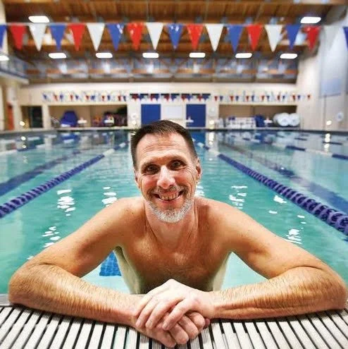Smiling man with short hair and facial hair leaning on the edge of an indoor swimming pool, with swimming lanes and flags overhead.