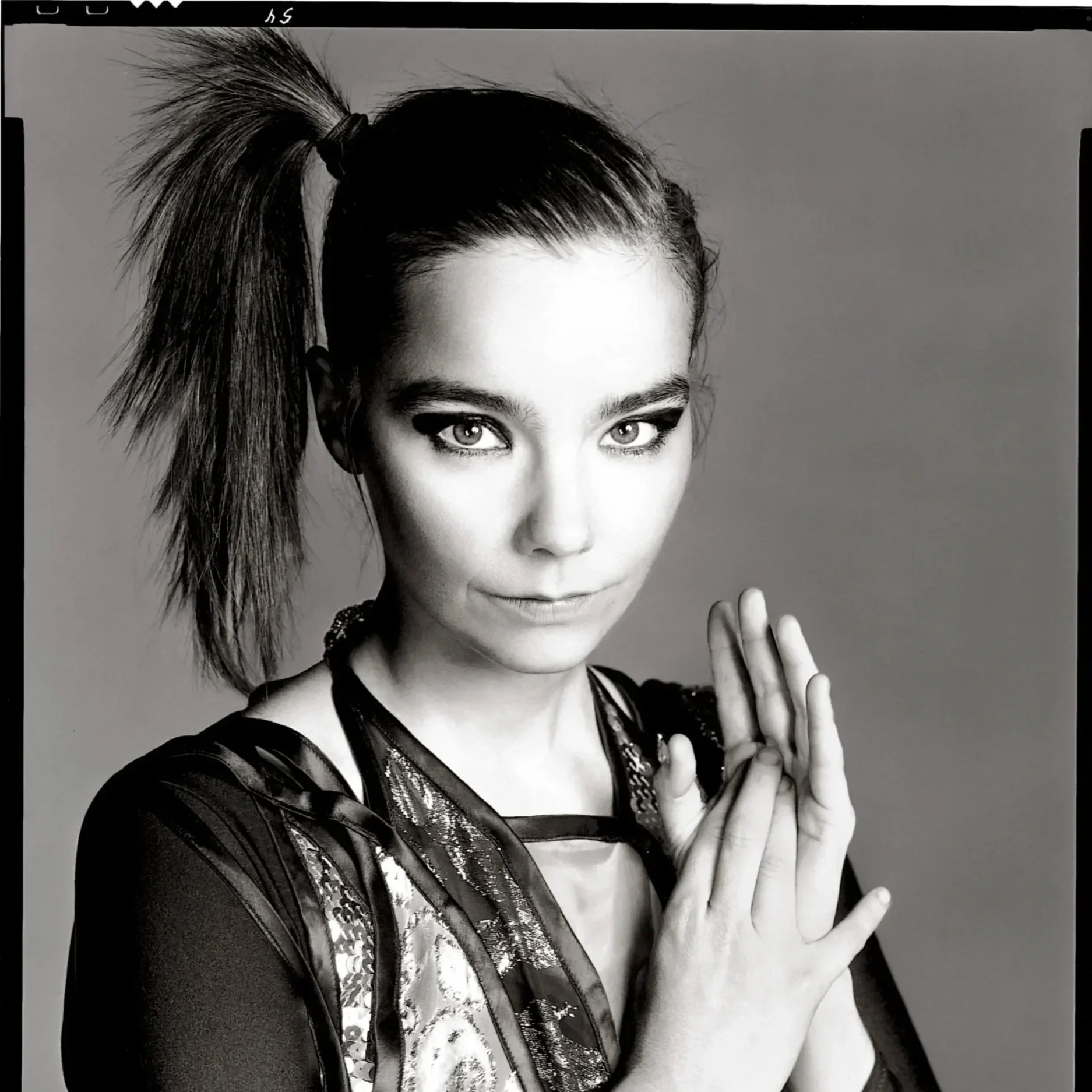 Black and white portrait of a woman with short hair styled upward and to the side, wearing a shiny top with intricate details, and making a gesture with her hands near her face.