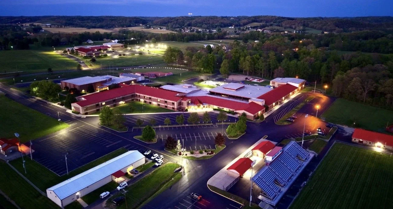 Aerial view of a large school complex at dusk, with multiple buildings illuminated, surrounded by parking lots and green fields.