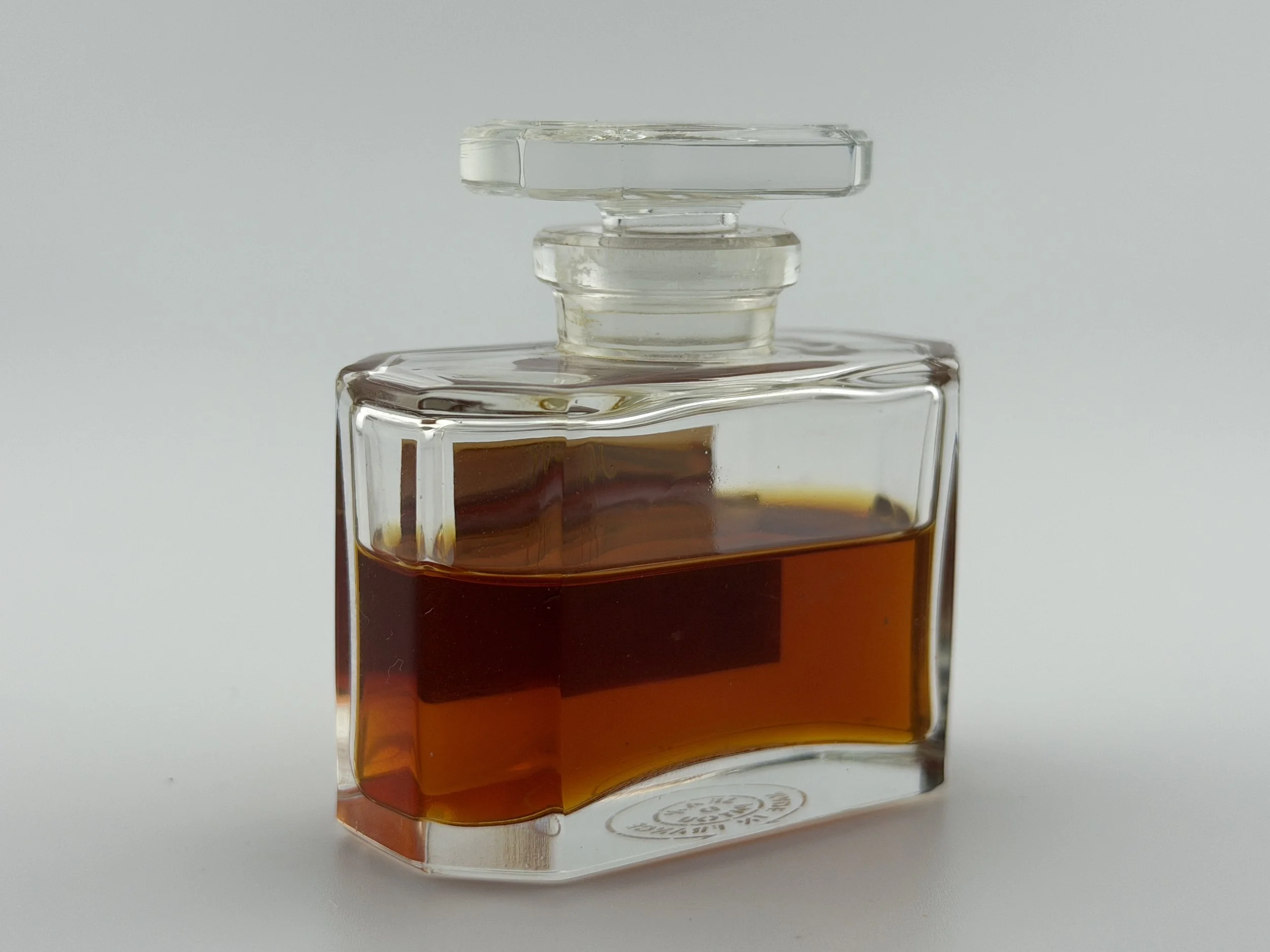 A valuable, sophisticated, vintage glass perfume bottle with a rectangular shape contains a dark amber liquid, with a glass stopper on top, set against a plain white background.