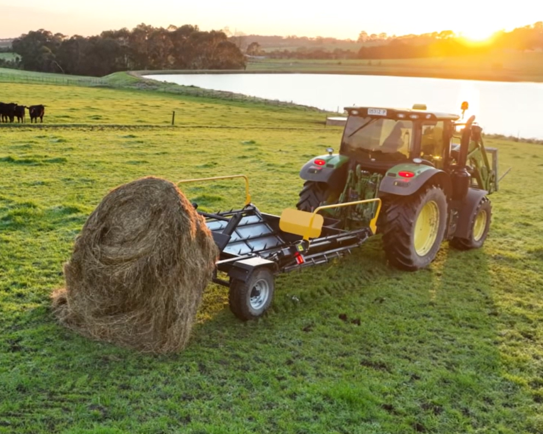 A tractor moving a large hay bale across a grassy field near a body of water at sunset.