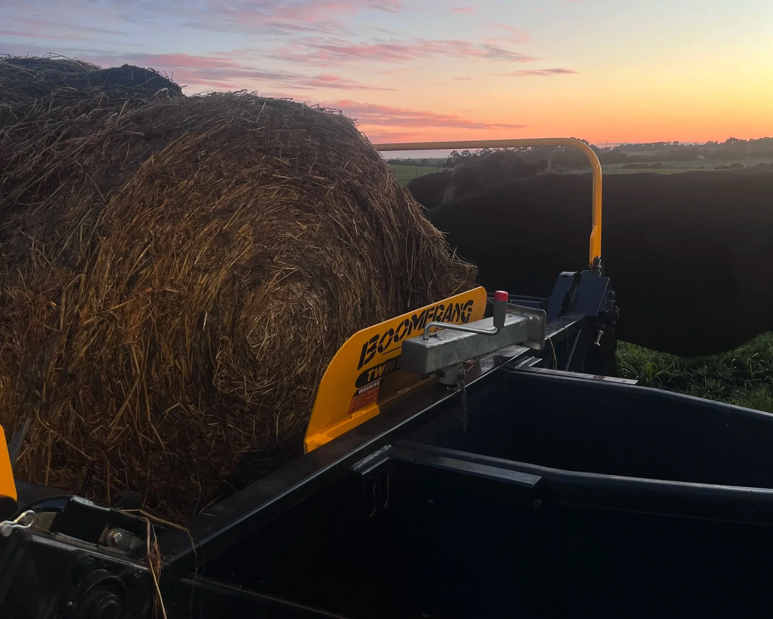 A hay baler machine on a farm during sunset, with a large round hay bale in the machine, open fields, and a colorful sky in the background.