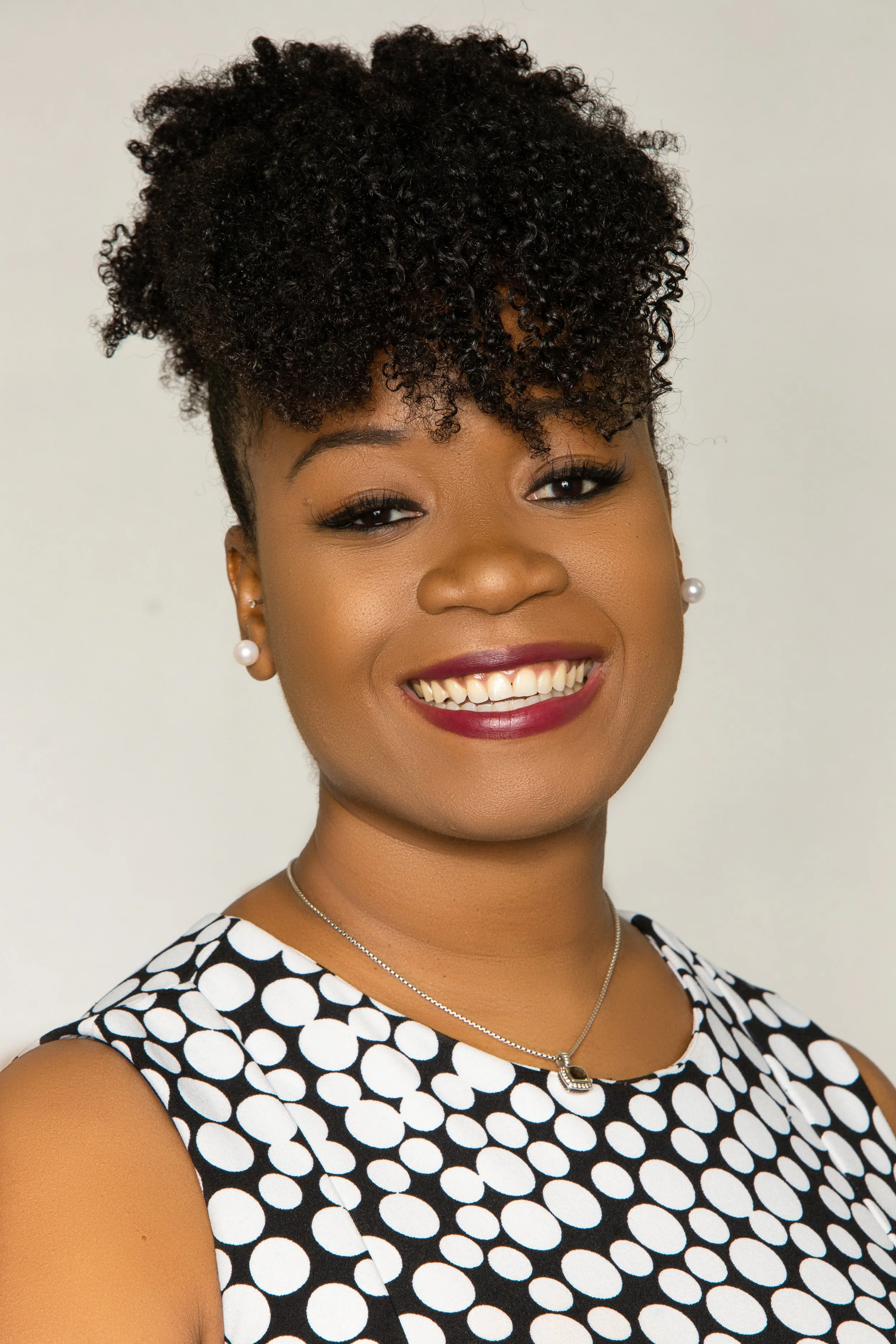 Close-up of a smiling Black woman with short, curly hair, wearing pearl earrings, a black and white polka dot dress, and a silver necklace with a pendant.