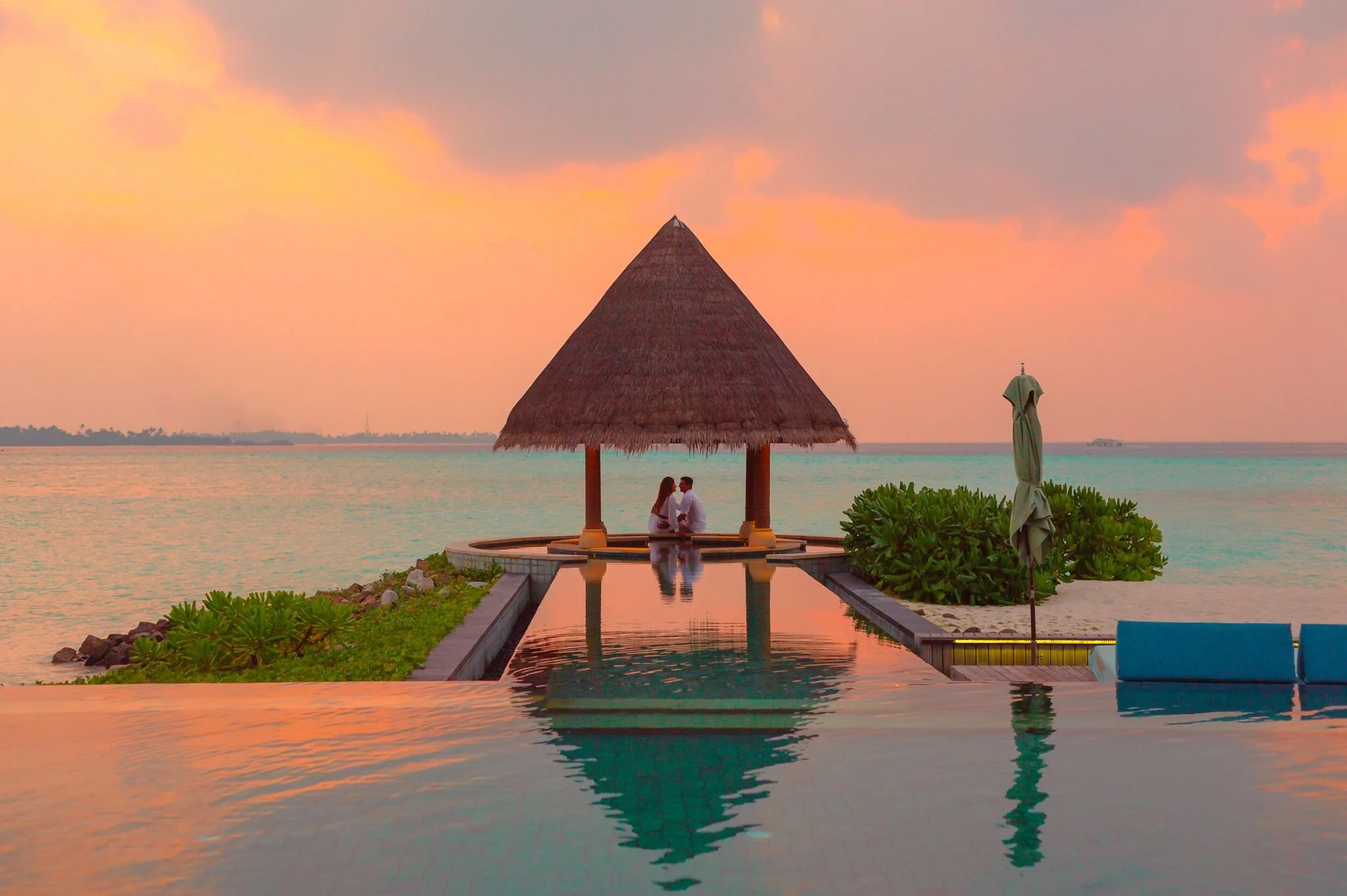 A couple sitting under a thatched roof hut on the beach at sunset, with the water, a small pool, and green plants surrounding them.