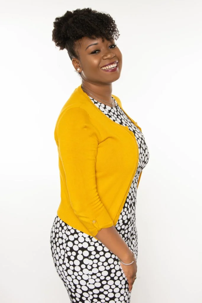 A smiling woman with curly hair tied back, wearing a yellow jacket and a black-and-white polka dot dress, standing against a plain white background.