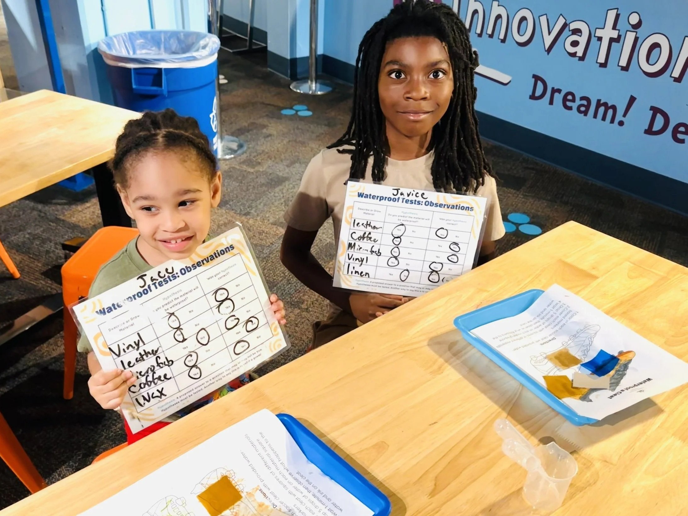 Two children sitting at a table, holding water test observation sheets with small circles and checkmarks, in a classroom or learning center with a blue water-related theme.