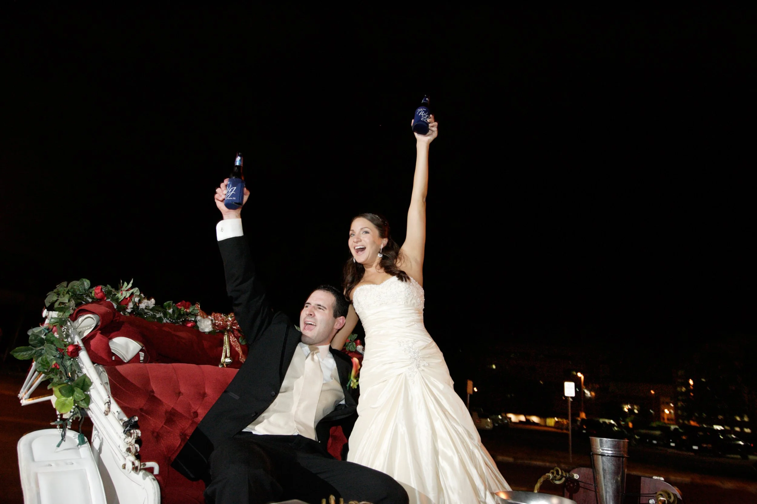 A newlywed couple riding in a decorated carriage during a nighttime celebration, with the bride in a white wedding dress and the groom in a black suit, holding drinks and smiling.