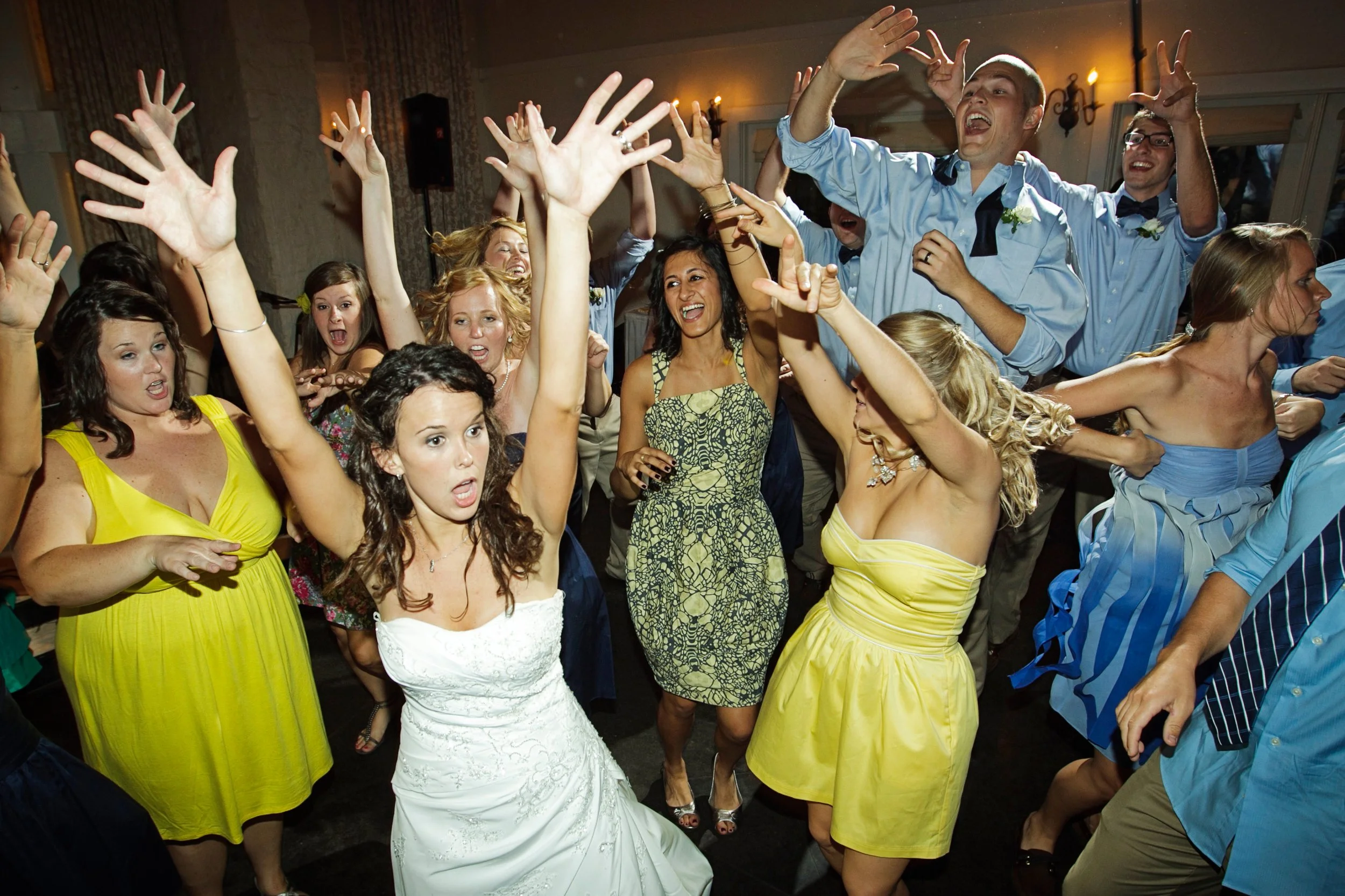 group of young adults dancing and celebrating at a wedding reception with some people raising their hands in the air and smiling.