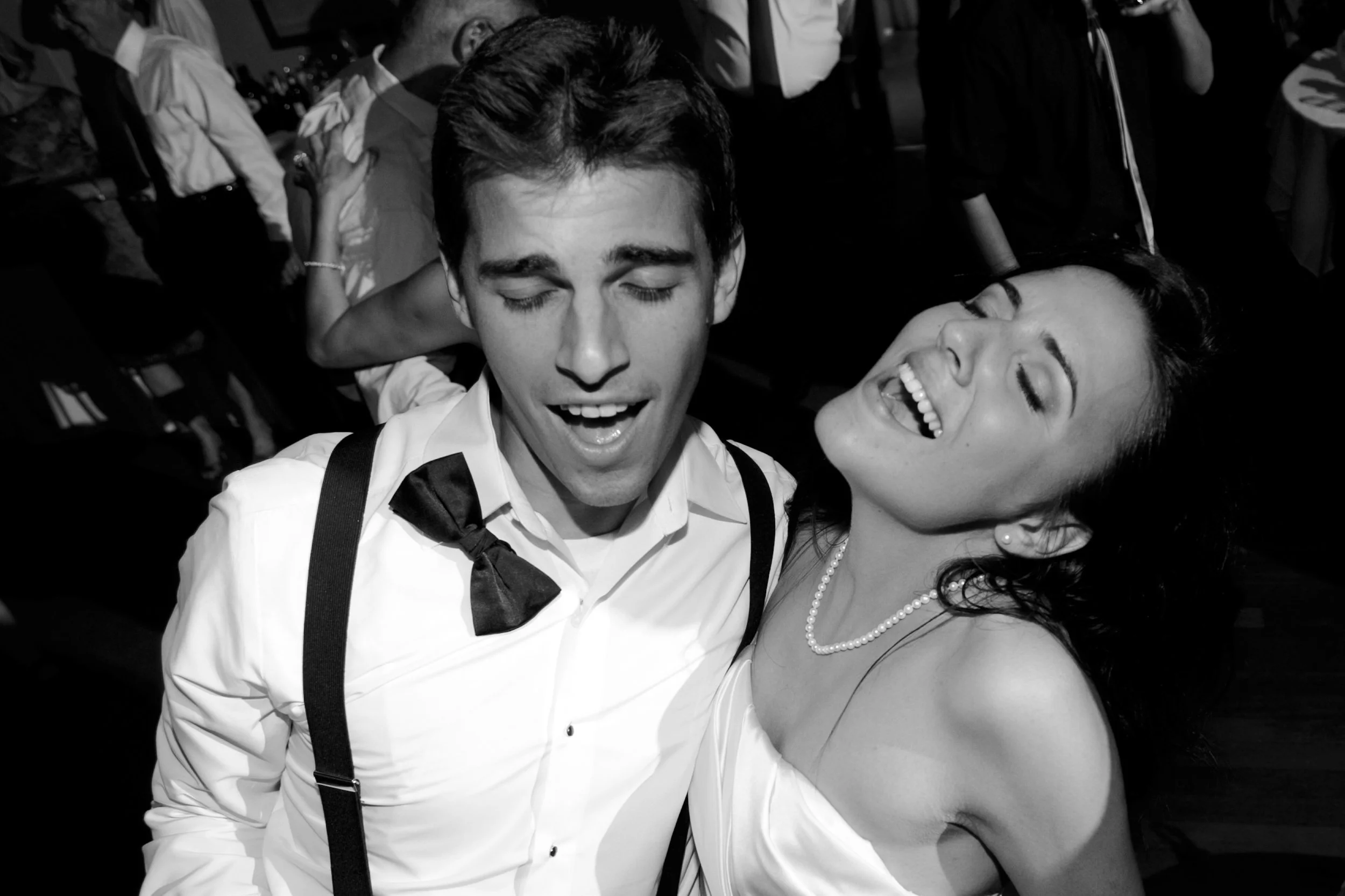 A black and white photo of a young man in a white shirt with suspenders and a bow tie, smiling with eyes closed, and a smiling woman with pearl jewelry, leaning close to him. They appear to be at a joyful social event or dance, with other people in t