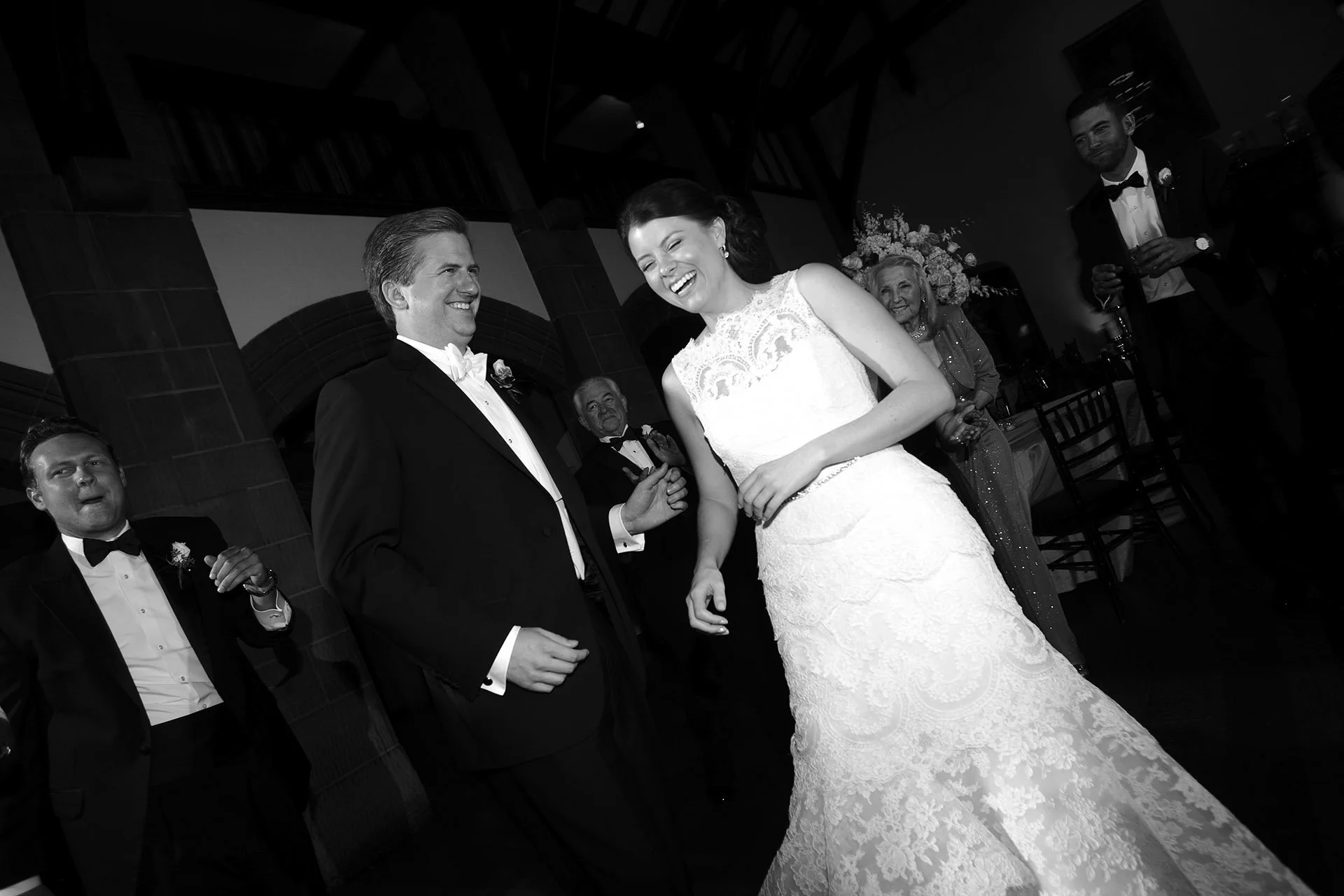 Black and white photo of a wedding reception, with a bride and groom smiling and holding hands, surrounded by guests in formal attire.