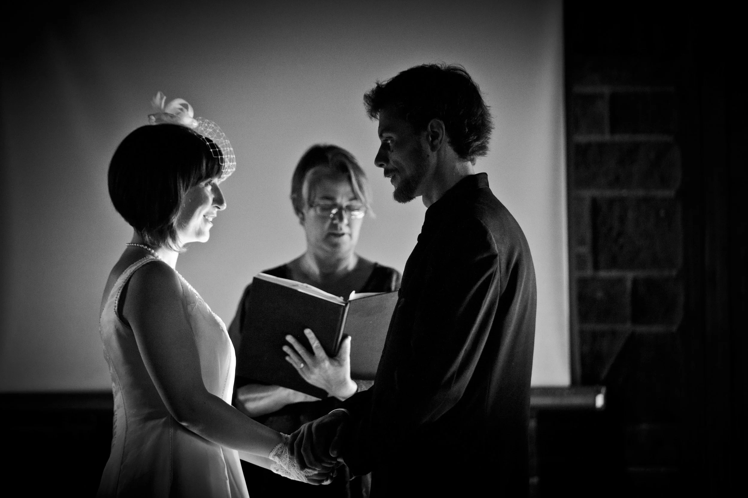 A black and white photo of a bride and groom exchanging vows, with an officiant reading from a book or script.