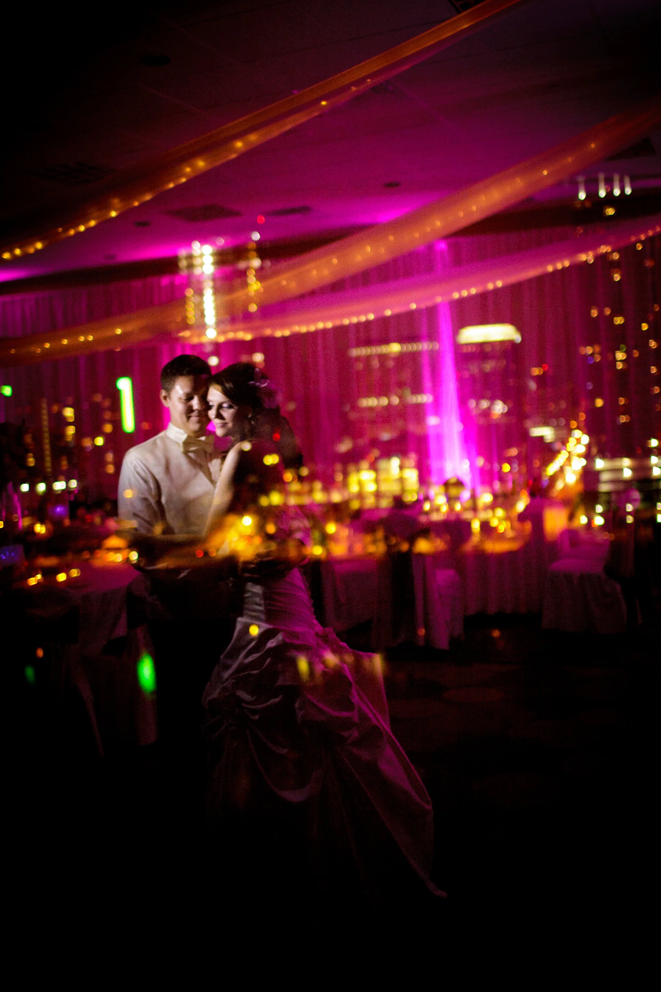 A couple dancing in a dimly lit venue with pink and purple lighting, city lights visible through the windows.
