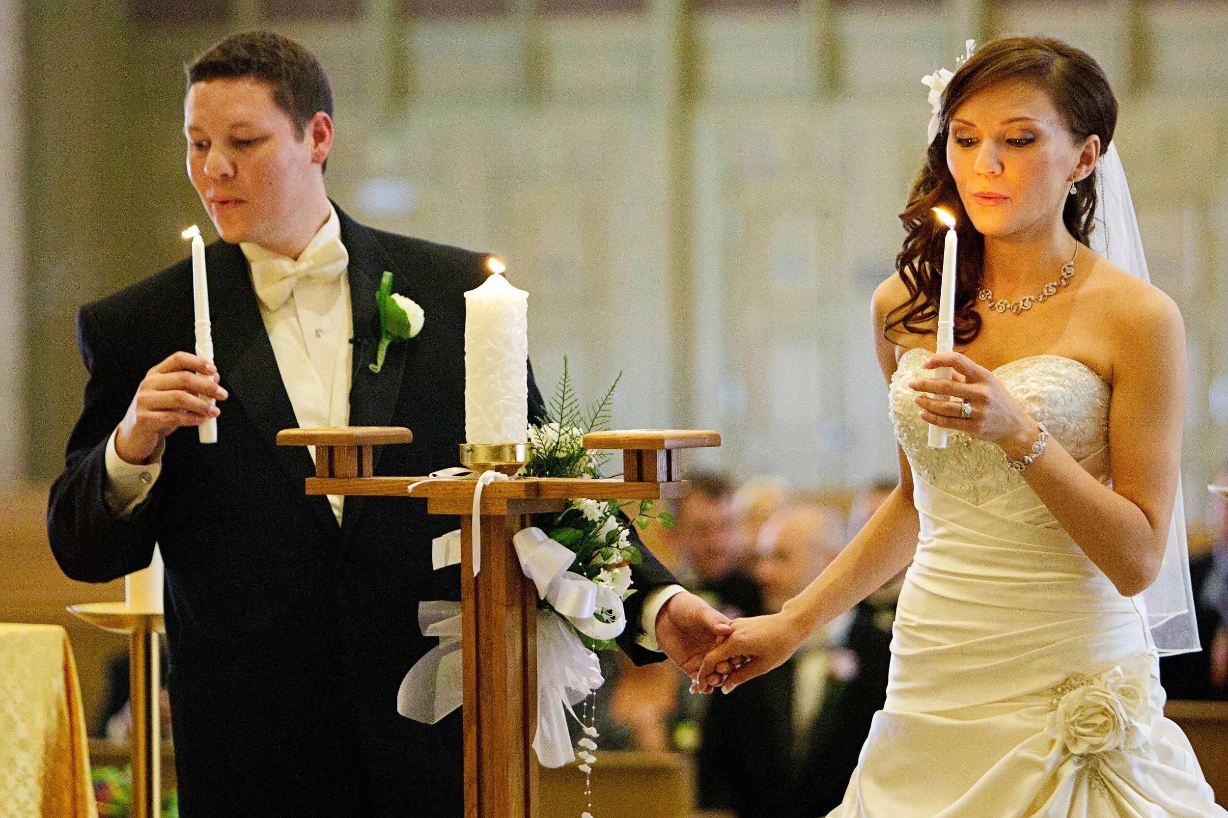 A newlywed couple holding hands during a wedding ceremony, with the bride wearing a white strapless wedding dress and the groom in a black tuxedo, both holding candles.
