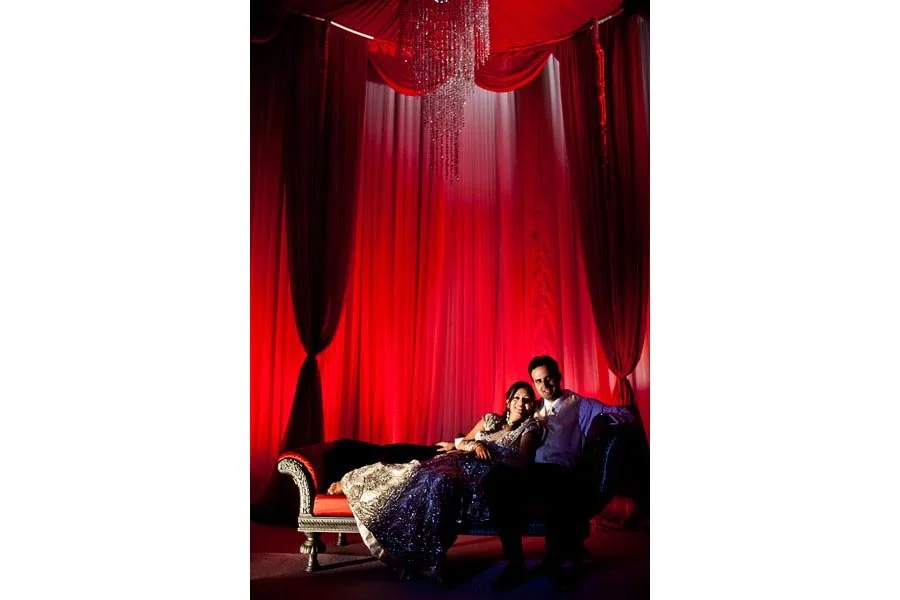 A couple sitting on a black and gold sofa in a room with red curtains and a chandelier, with a red and purple backdrop.