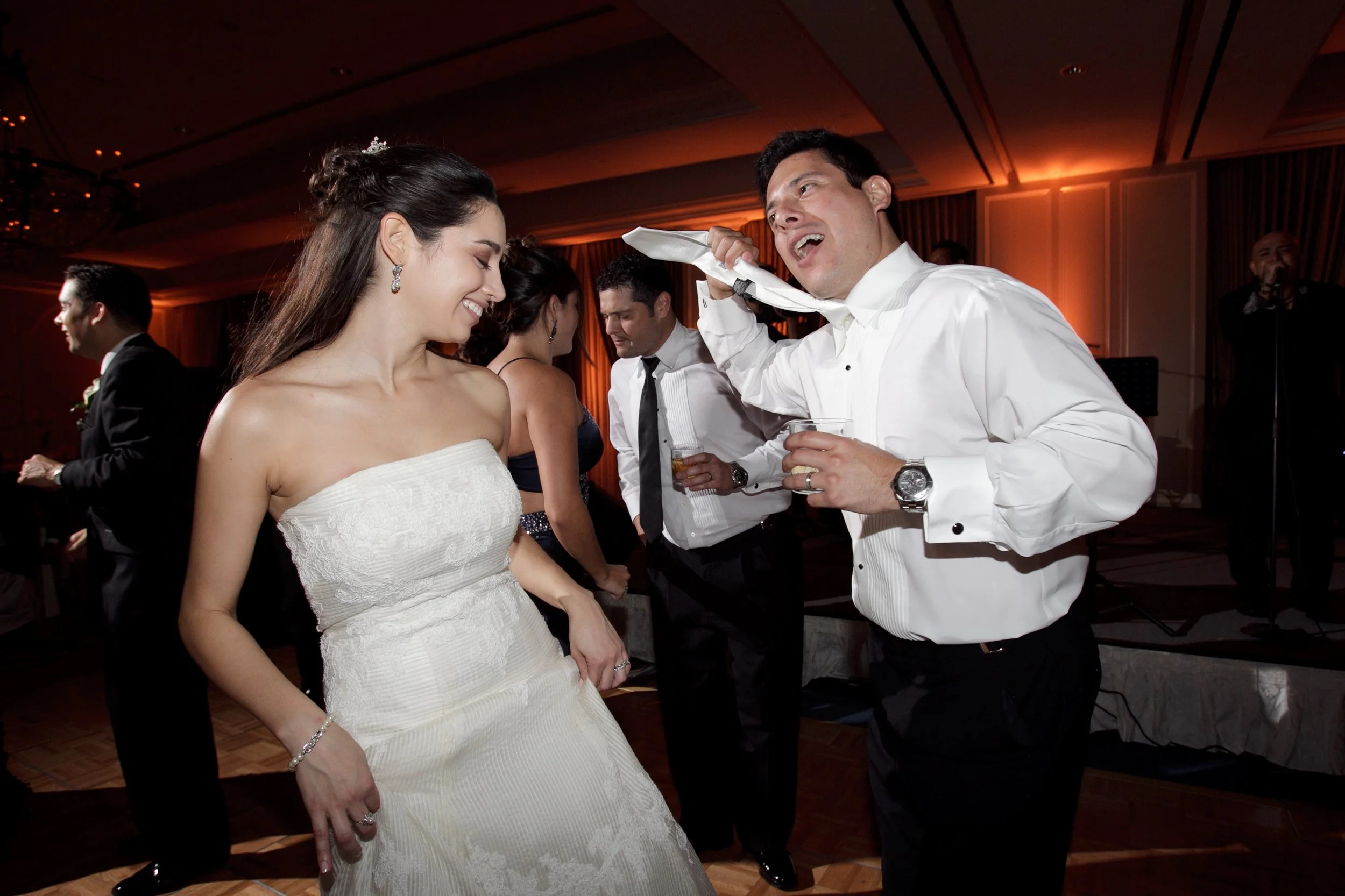 Bride in a wedding dress dancing and smiling with a man in a white shirt at a lively indoor celebration, with other guests in the background.