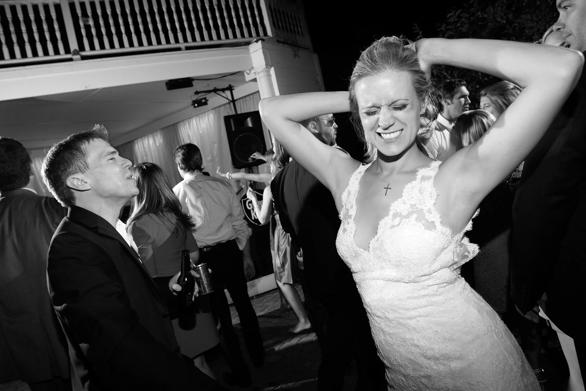 A woman in a wedding dress with her eyes closed and hands behind her head dancing at a celebration with people in the background.
