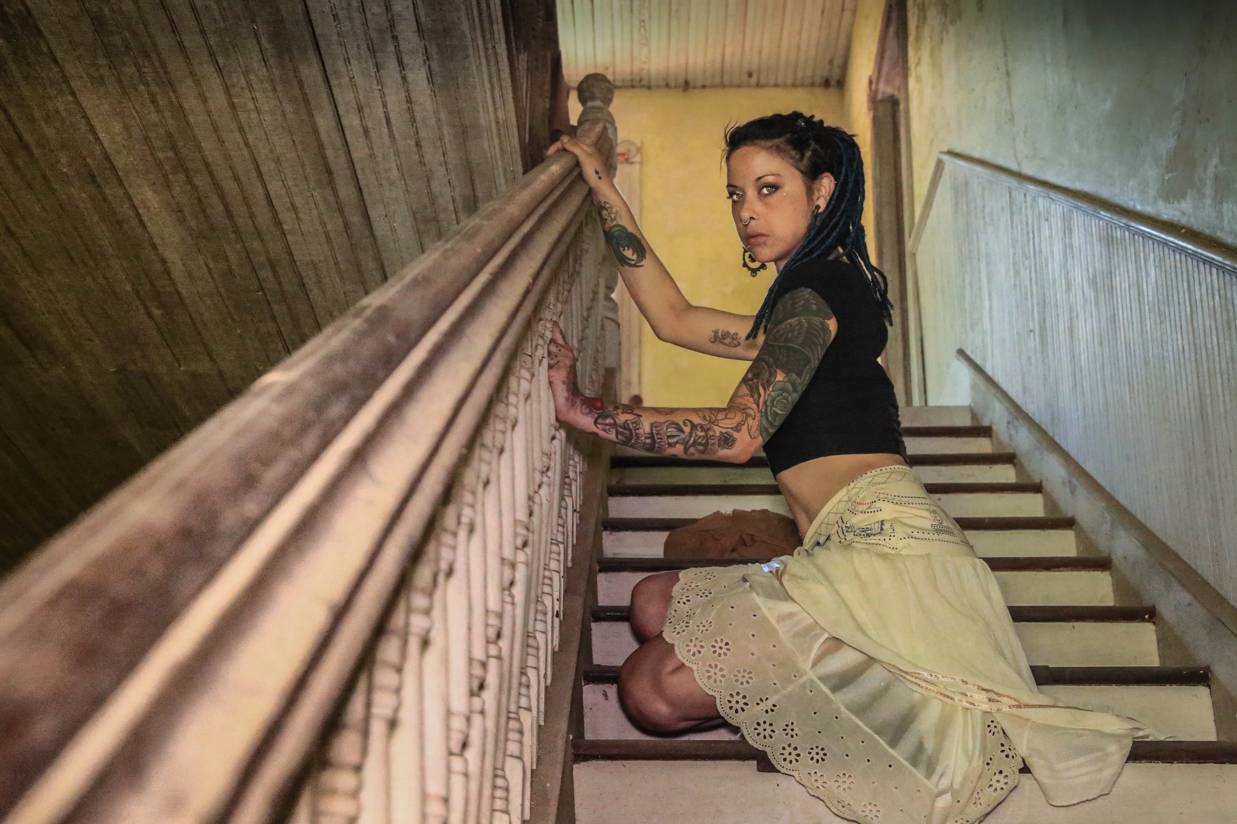 A woman with tattoos and dark braided hair is kneeling on a staircase, holding onto the railing, with another person in yellow shorts and a lace skirt visible in the foreground.