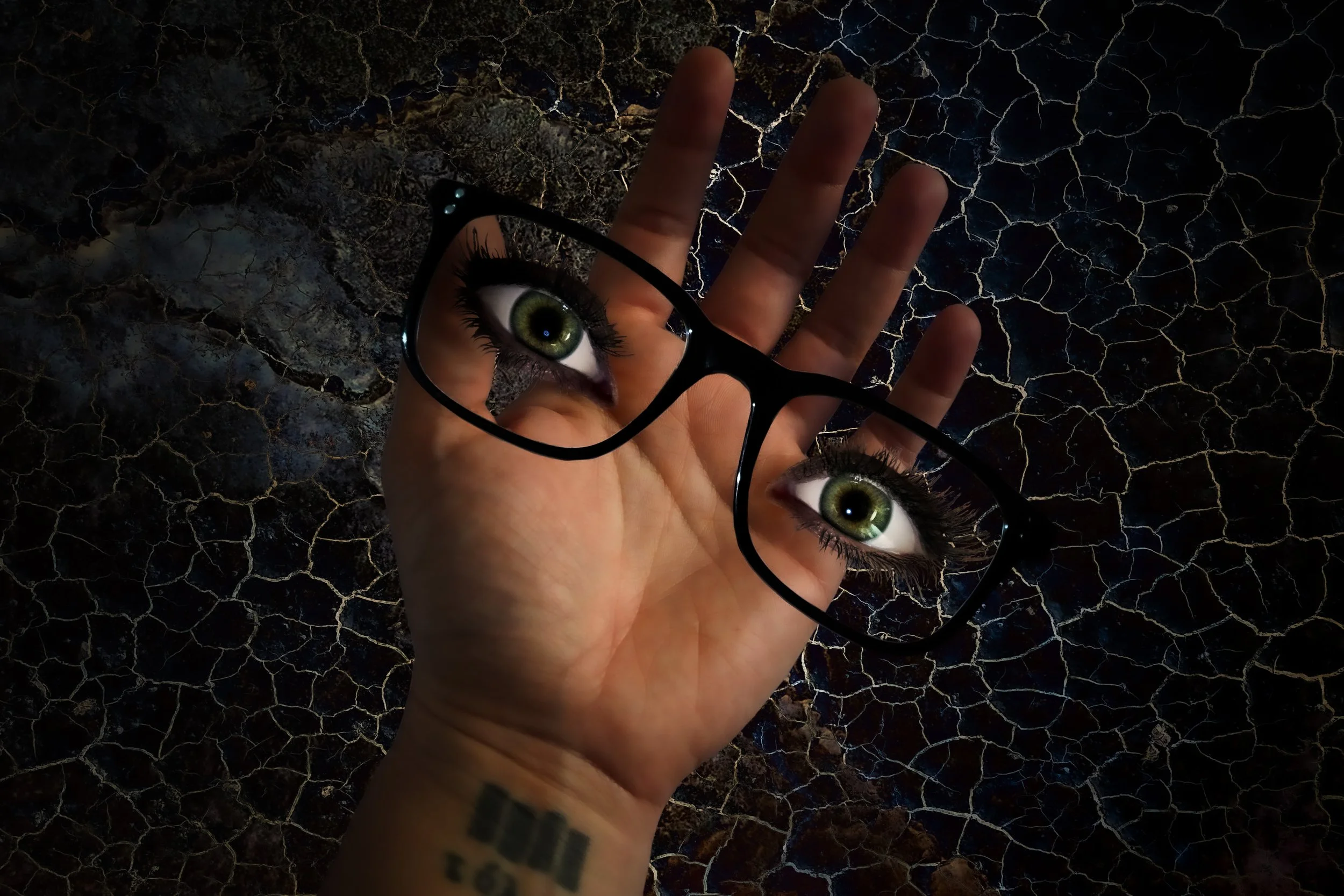A hand holding a pair of glasses with lenses that have eye designs in them, against a dark background with a cracked, textured surface.