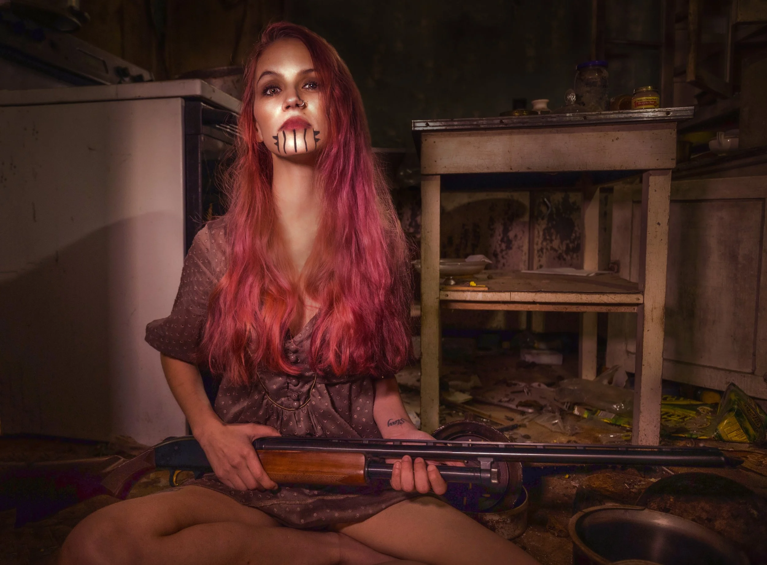 Young woman with red hair, face painted with black lines, holding a rifle in a dimly lit rustic kitchen with old furniture and scattered objects.