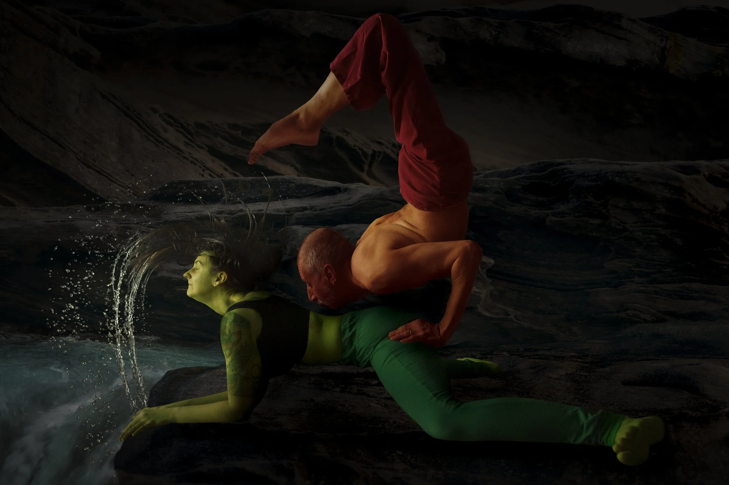 A person with green skin and patterned green clothing practicing yoga on rocks near water, with another person in the background doing a headstand on rocks.