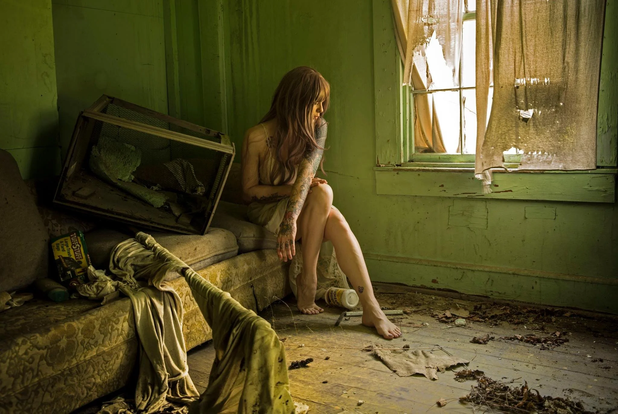 A woman with long hair and tattoos sitting on a worn, green wall in an abandoned room with torn curtains, debris on the floor, and outdated furniture.