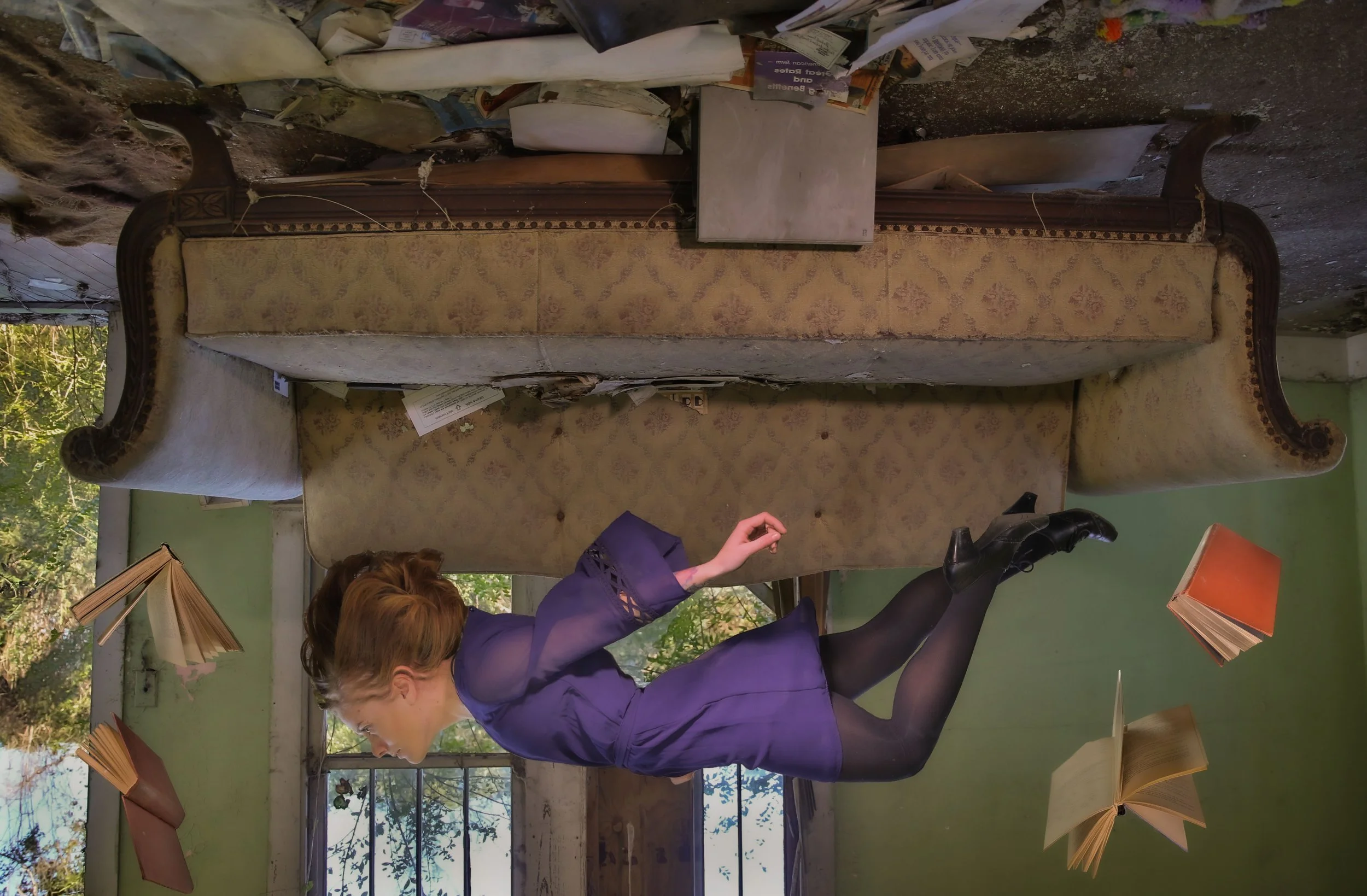 Woman in purple blouse sitting at a desk with open books, with a large vintage sofa behind her and a wall with windows showing trees outside.