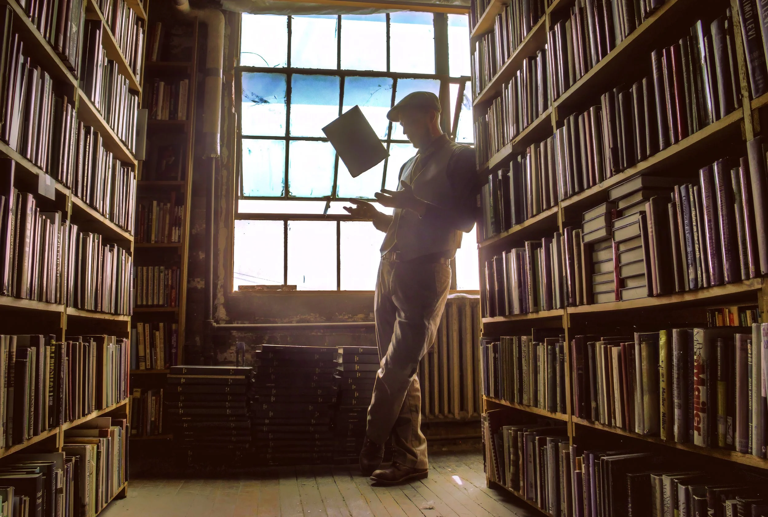 Person standing between bookshelves in a library, looking at a book, backlit by a large window with divided panes.