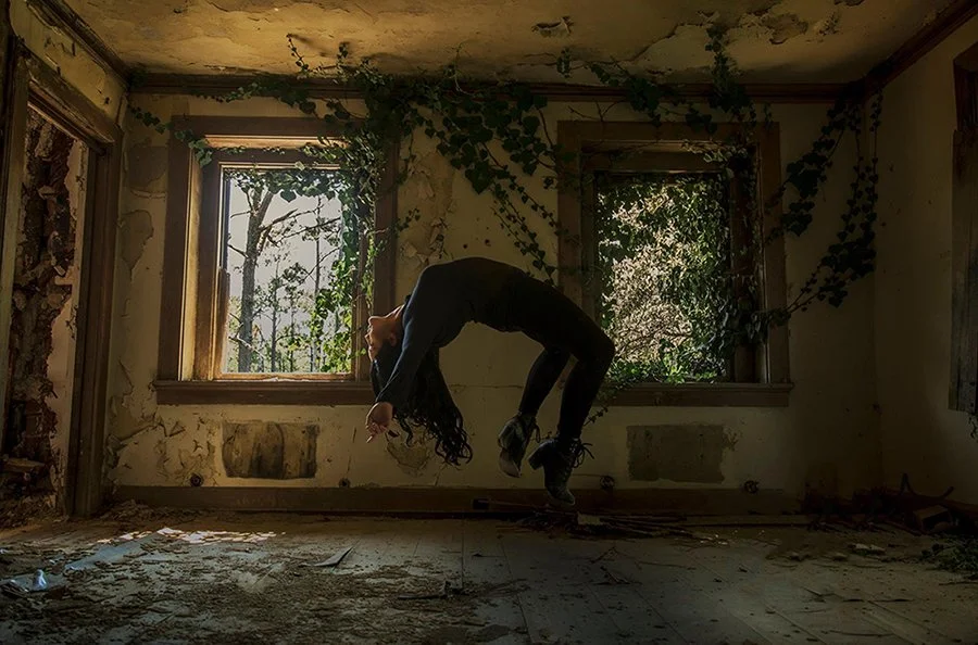 A woman with long dark hair is performing a backbend inside an abandoned, dilapidated room with peeling walls and overgrown plants coming through the windows.
