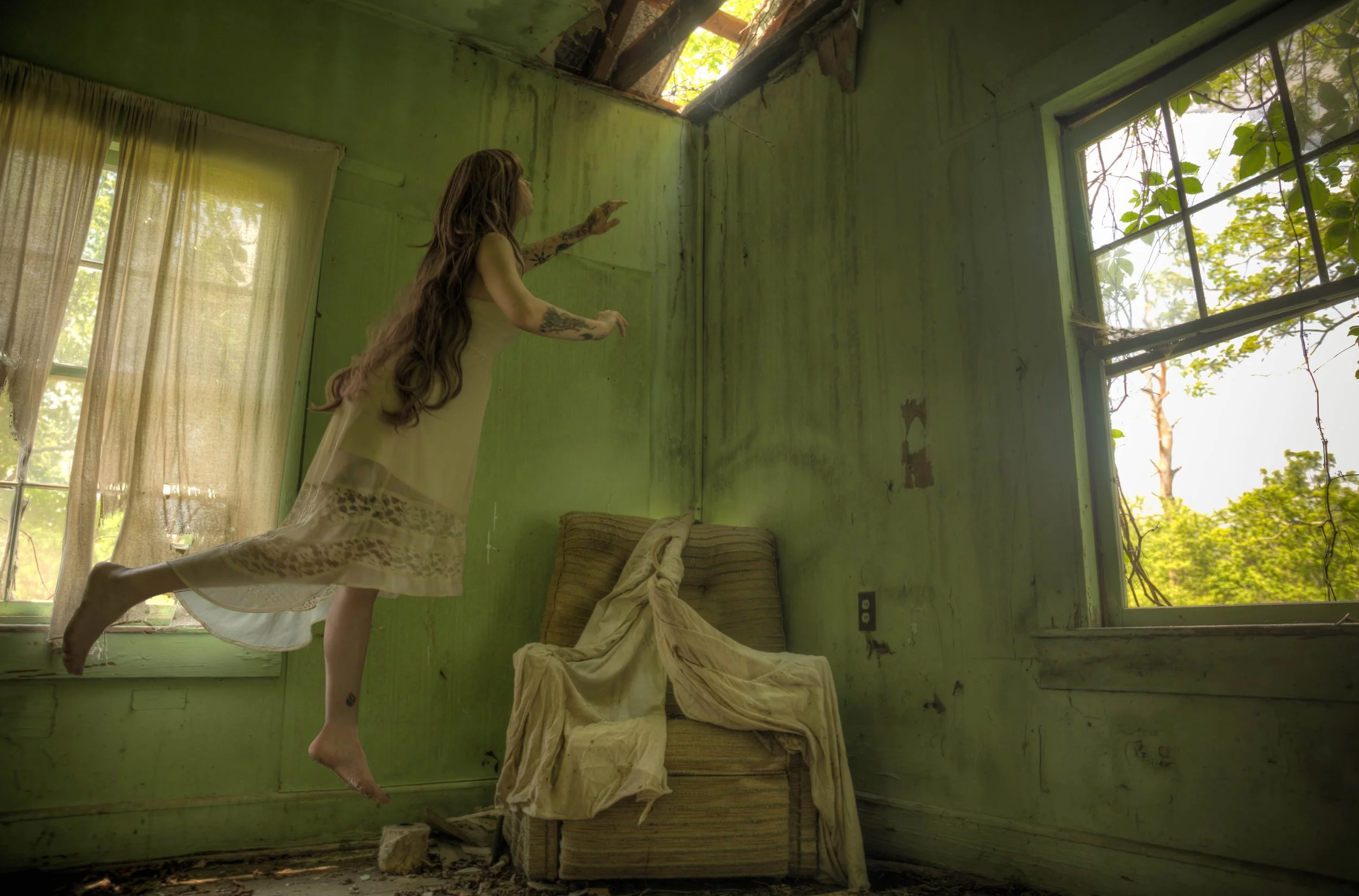 A woman with long hair and tattoos wearing a sleeveless dress is floating or walking on the ceiling of an old, abandoned room with green walls, large windows, and overgrown vegetation visible outside.