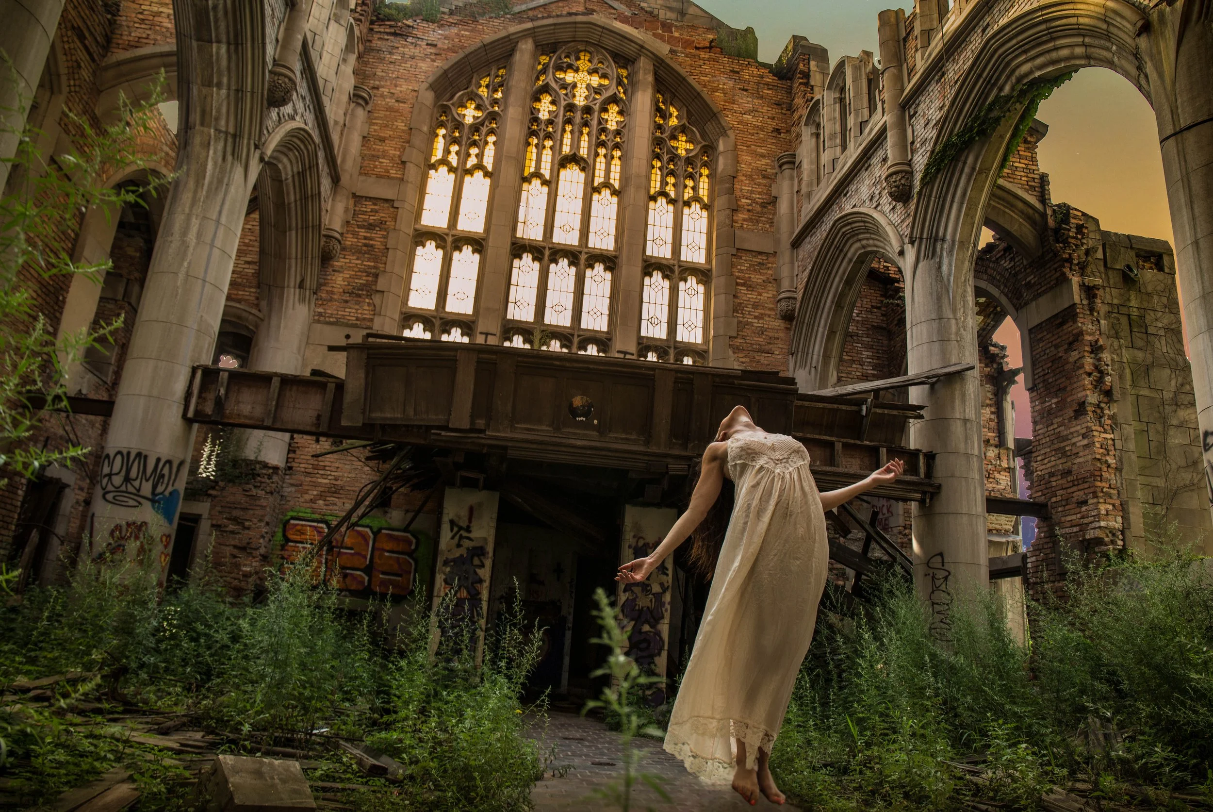A person wearing a long, light-colored dress is dancing or performing in front of the ruins of an old building with large stained-glass windows and archways. The building appears to be abandoned and partially overgrown with vegetation, with graffiti on some walls and graffiti on a pillar to the left. The scene is set during sunset or dusk, creating a warm glow in the sky.