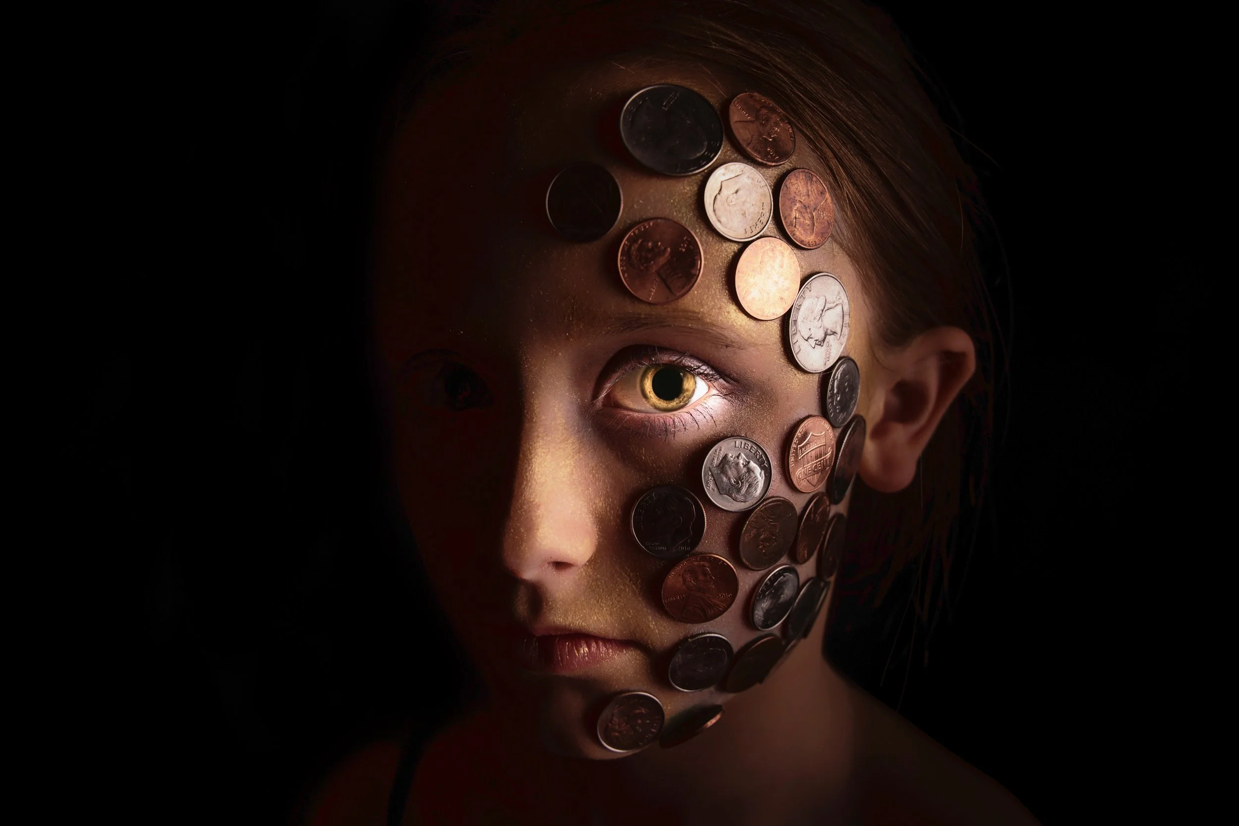 A person's face partially visible with only the eye showing, covered with various coins from different denominations and countries, set against a dark background.