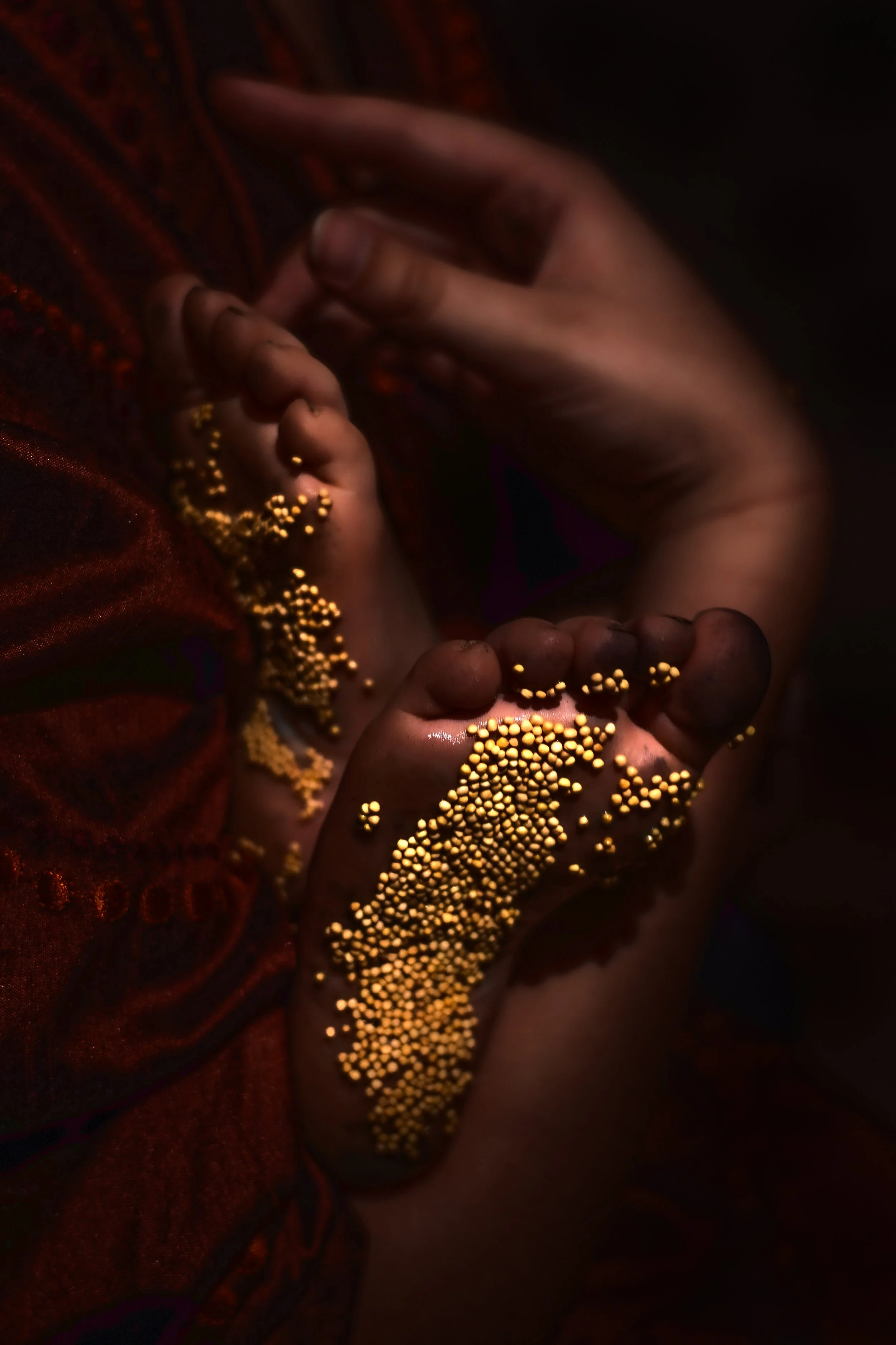 A close-up of hands holding tiny yellow beads, with some beads spilling onto the fingers and a dark red fabric background.