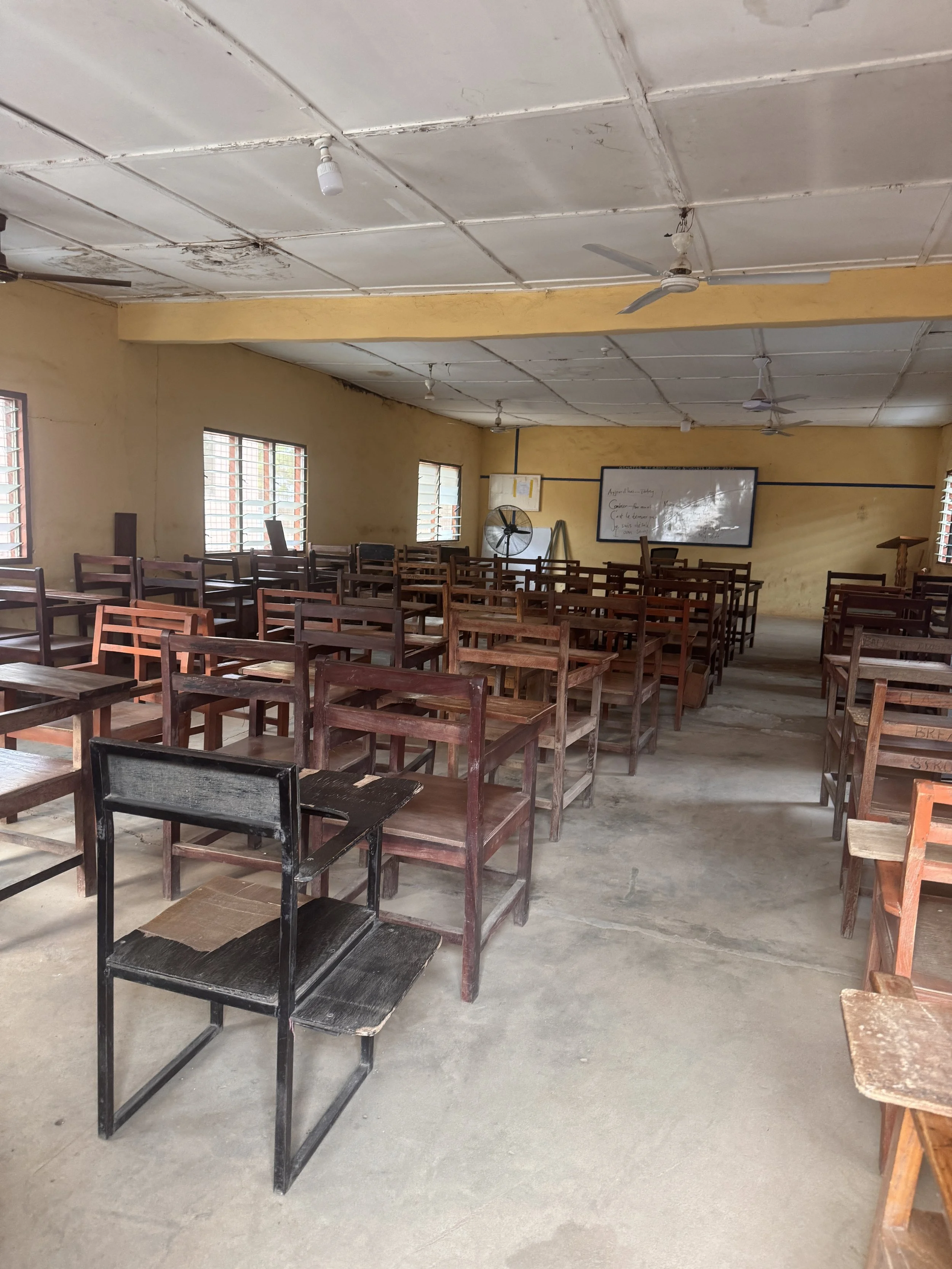 An empty classroom with wooden desks and chairs, yellow walls, large windows with metal bars, a whiteboard, ceiling fans, ceiling lights, and a concrete floor.
