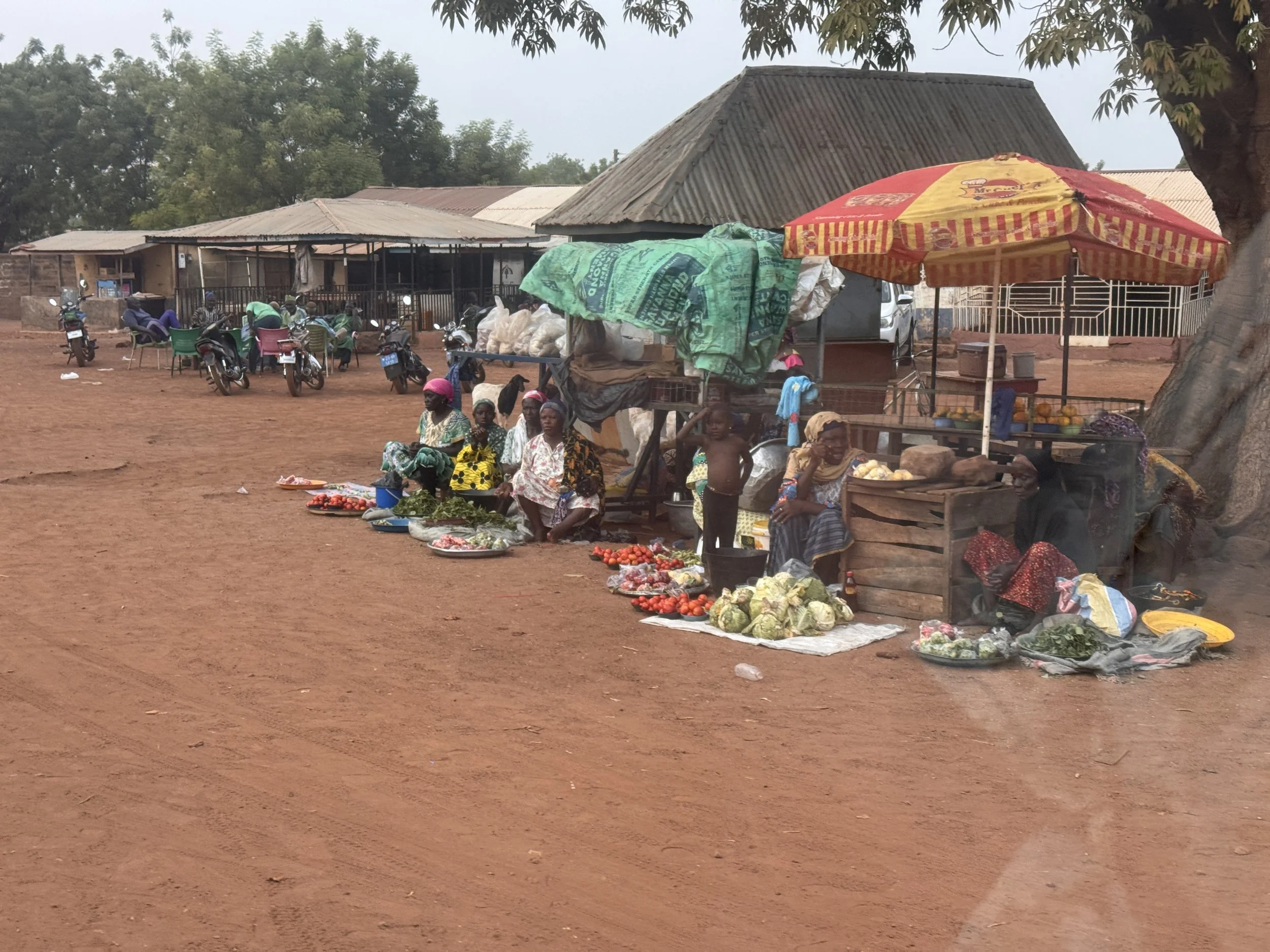 A rural market scene with women selling vegetables, including tomatoes, on the ground and in baskets, with a colorful umbrella providing shade; several motorcycles are parked nearby, and there are people sitting and walking on reddish dirt ground, with simple buildings and trees in the background.