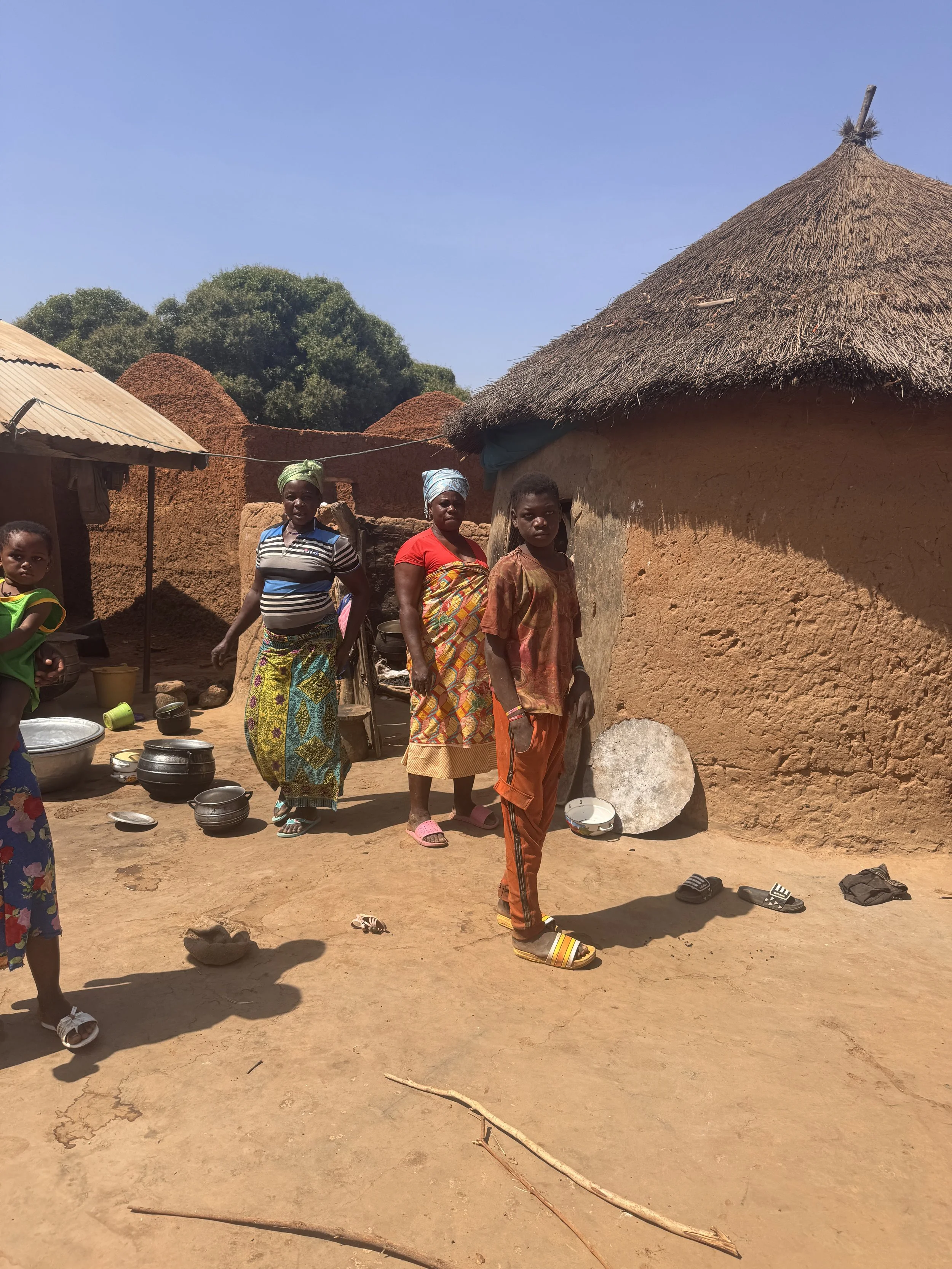 A group of five women and children standing outside traditional mud and thatch huts in a rural village. The ground is dirt, and household items such as bowls and sandals are visible.