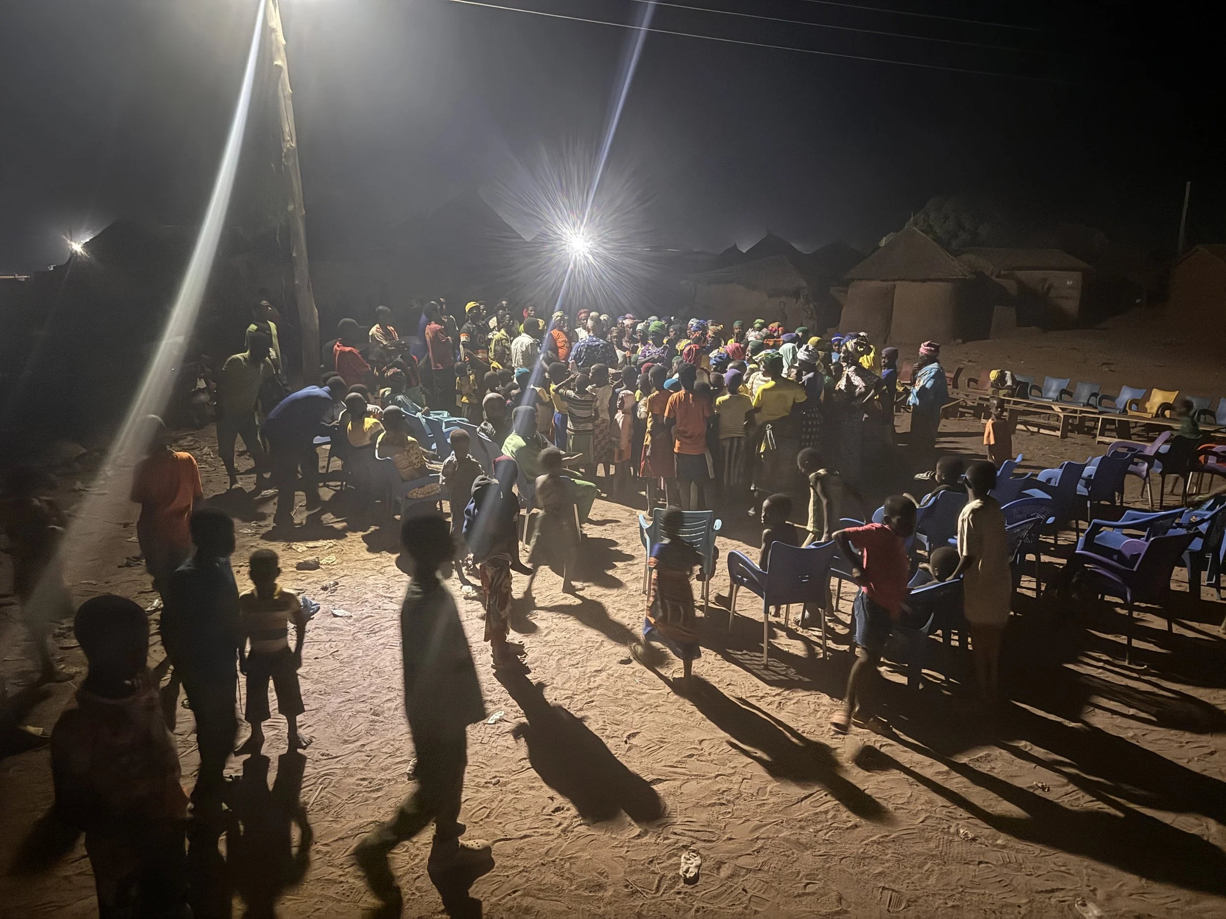 A large crowd of people gathered outdoors at night, some standing and some sitting on chairs, with a bright light source illuminating the scene.