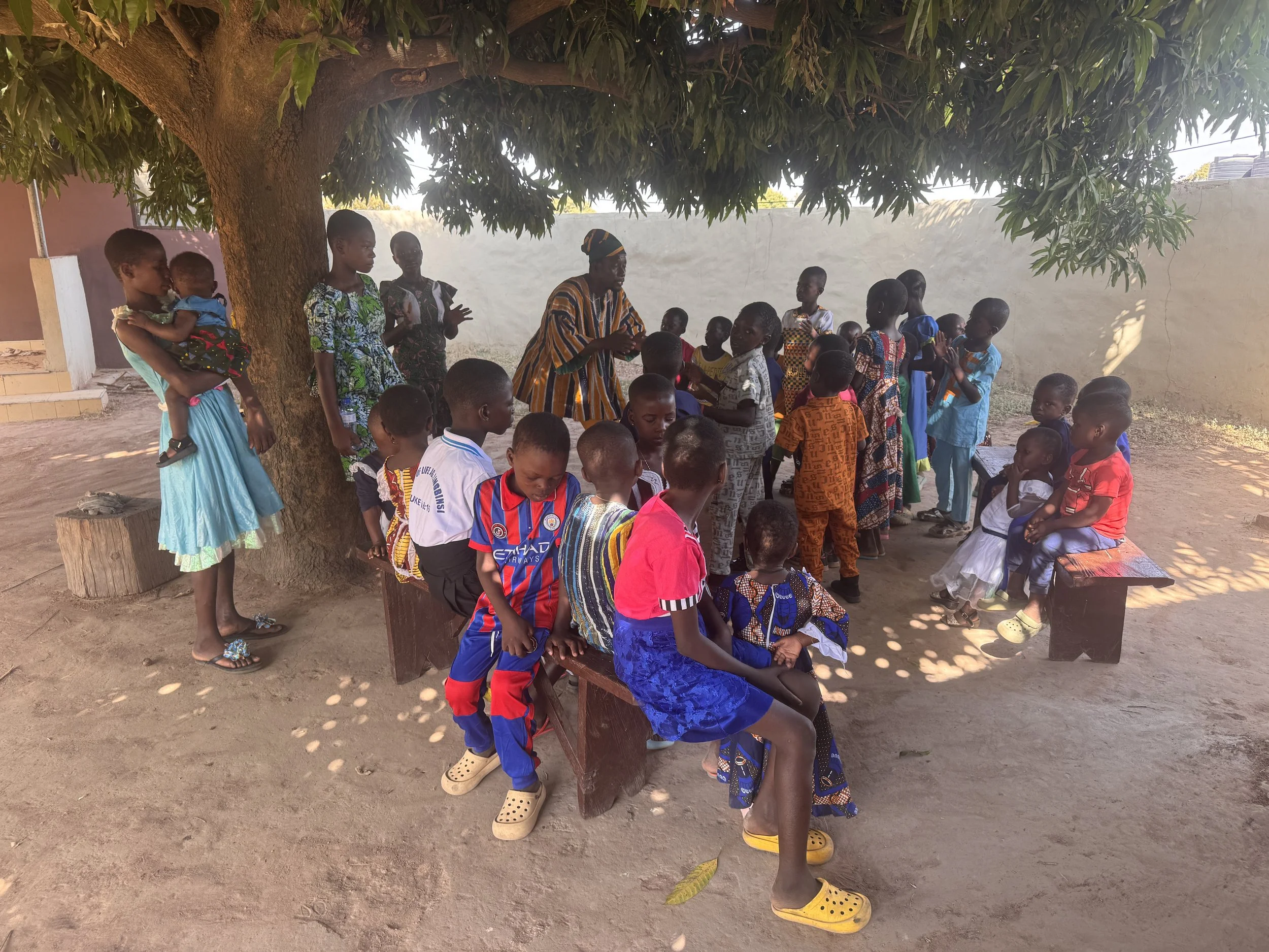 A group of children and a woman gathered under a large tree in an outdoor setting, some sitting and some standing, with a plain wall in the background.