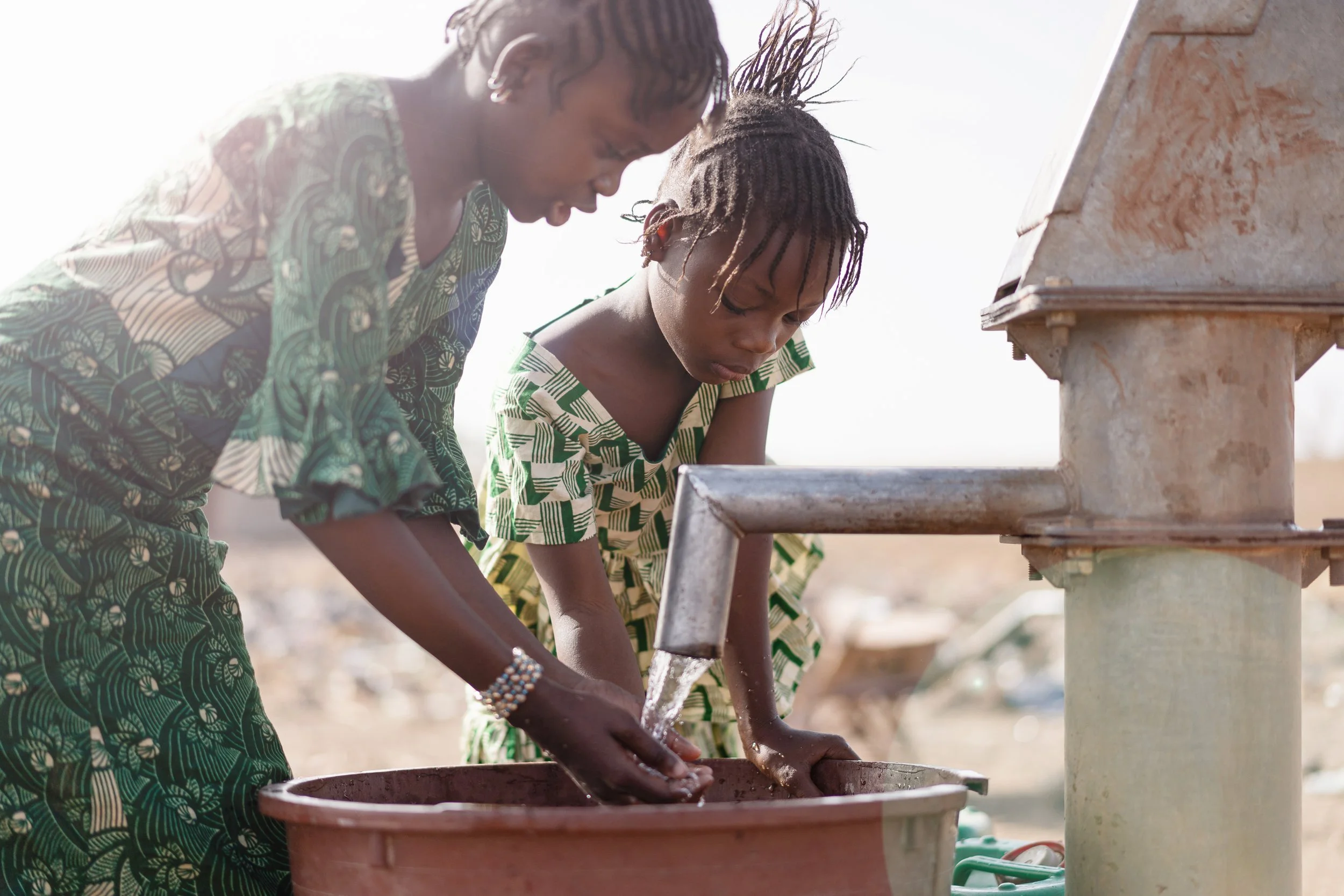 Two young women with African features washing their hands at outdoor water station, wearing patterned dresses, in a bright outdoor setting.